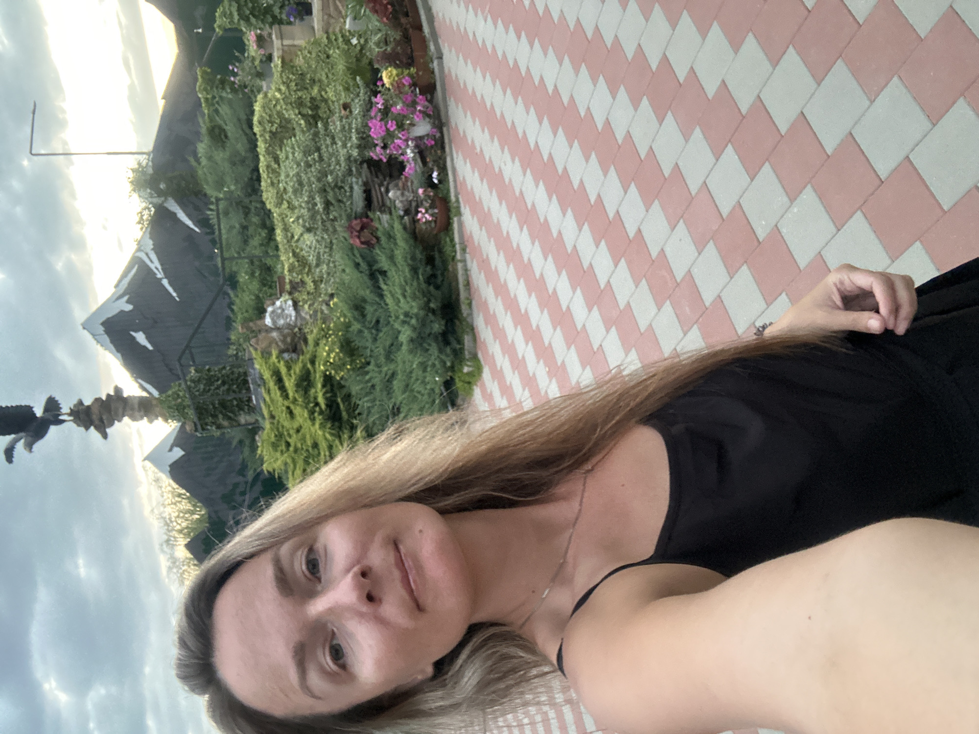 A woman with long blonde hair takes a selfie in front of a decorative garden scene. She is wearing a black tank top and a silver necklace. The background features artificial mountains, lush green bushes, and pink and white flowers in planters. A statue of an eagle perched on a rock column stands prominently in the upper left background. The ground is paved with a diamond pattern of pink and light gray tiles. The sky above is cloudy with hints of sunset light. The location is described as being in the city of Yaroslavskaya, Russia.