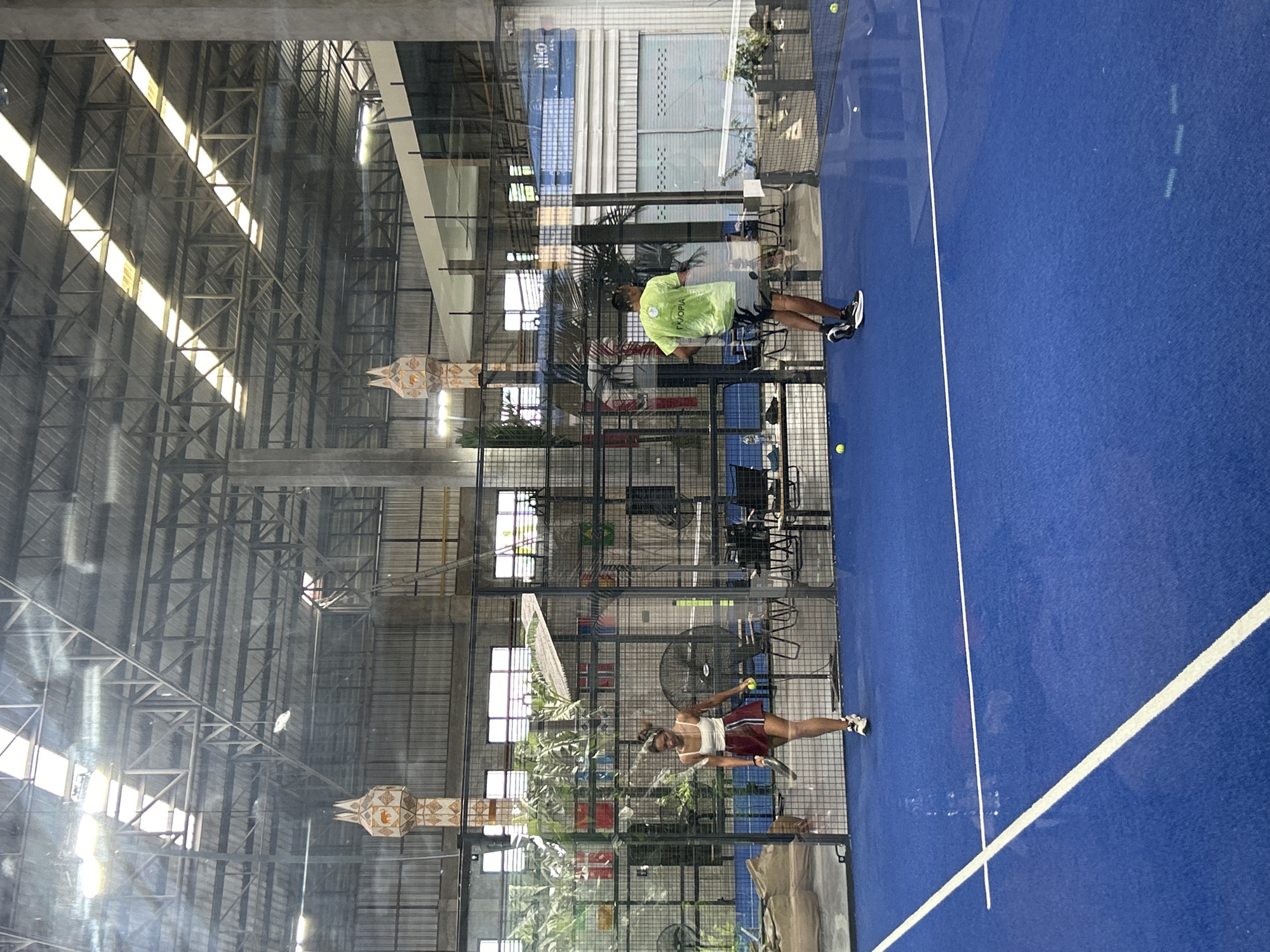 The image depicts an indoor padel court with a bright blue surface and white boundary lines. The court is enclosed by glass walls and metal fencing, with a high ceiling supported by industrial metal beams. Natural light streams in from windows along the side of the building, creating bright stripes of light on the court and illuminating the metal structure.
Two people are visible on the court, engaged in a game of padel. One is a woman wearing a white tank top and a maroon skirt, holding a padel racket and a ball, appearing to be in the process of serving or hitting the ball. The other person is a man in a lime green t-shirt and dark shorts, standing on the other side of the court. He is facing away from the camera, his body in a stance that suggests he is watching the game.
Decorations hang from the ceiling, including what appear to be traditional paper lanterns with ornate patterns. Plants and greenery are visible behind the glass walls, suggesting an outdoor or semi-outdoor area beyond the court. Fans are mounted on the walls inside the enclosed area.
The overall lighting suggests it is daytime. The scene appears to be in an indoor sports facility, likely in a warm climate given the presence of lush plants. No overt emotions are discernible from the subjects, but their postures indicate active participation in a sport. The text "ΓΚΟΡΙΑ" is visible on the man's t-shirt. The location context places this scene in Ban Buak Khrok North, Thailand.