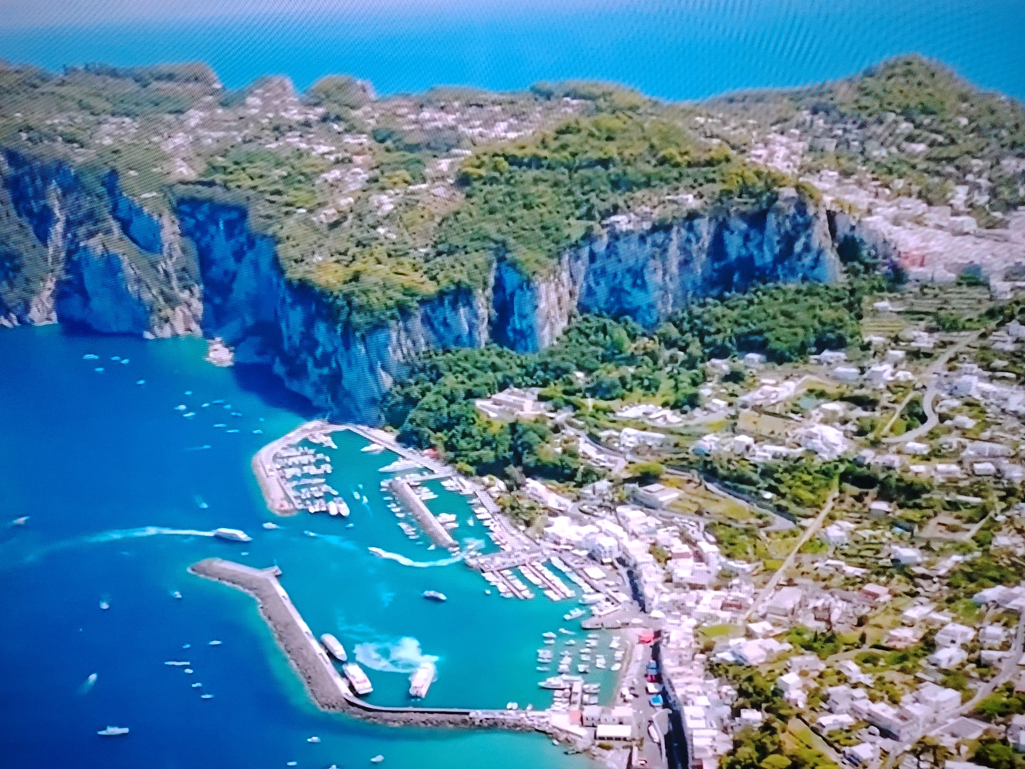This is an aerial, high-angle, slightly blurry, digitized photograph of a coastal town, likely situated on a Mediterranean island. The foreground features a vibrant turquoise harbor filled with numerous boats of various sizes, many docked along piers. The water exhibits some turbulence, suggesting the movement of vessels, with white wakes visible. A breakwater extends from the lower right of the frame, creating a protective barrier for the harbor.
The middle ground reveals a densely populated town characterized by white buildings with terracotta roofs, nestled against a steep, verdant hillside. The town extends up the slopes, with winding roads and pathways visible. Lush green vegetation covers the upper slopes of the hills, creating a striking contrast with the white buildings. In the background, sheer, rocky cliffs, partially covered in greenery, drop dramatically towards the sea. The sky is a clear, bright blue.
There are no discernible people or specific identifiable landmarks that would indicate the location as Huntingdonshire, United Kingdom. The architecture and landscape are distinctly Mediterranean.