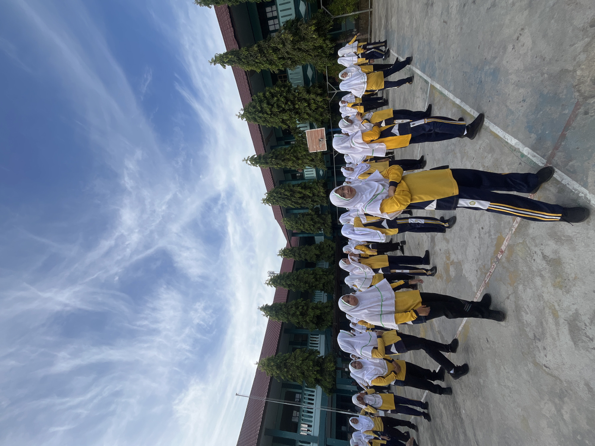 This image depicts a group of female students wearing yellow long-sleeved shirts, dark pants with white and yellow stripes, and white hijabs, standing in an outdoor courtyard. The students appear to be participating in an organized activity, possibly a school event or physical education class.
The location is the courtyard of a school building, characterized by its blue and green facade, red roof, and tall green trees lining the perimeter. The ground is paved with concrete, marked with white lines, suggesting it might be a basketball court. A basketball hoop with a wooden backboard is visible in the background.
The sky is bright blue with scattered white, wispy clouds. The overall lighting suggests it is daytime, likely morning or afternoon. The students are facing towards the right side of the frame, with some looking towards the camera. Their postures are varied, indicating they are either standing at attention or in the process of moving.
The scene is set in the city of Lhokseumawe, Indonesia, which is known for its tropical climate and educational institutions. The image captures a moment of daily life at a school, highlighting the students' uniforms and the typical environment of an Indonesian educational facility. The forensic aspect of the image is its objective documentation of the scene, capturing the details of the individuals, their attire, the architecture of the school, and the natural elements present. There are no obvious signs of conflict or distress, suggesting a peaceful and ordinary occurrence.