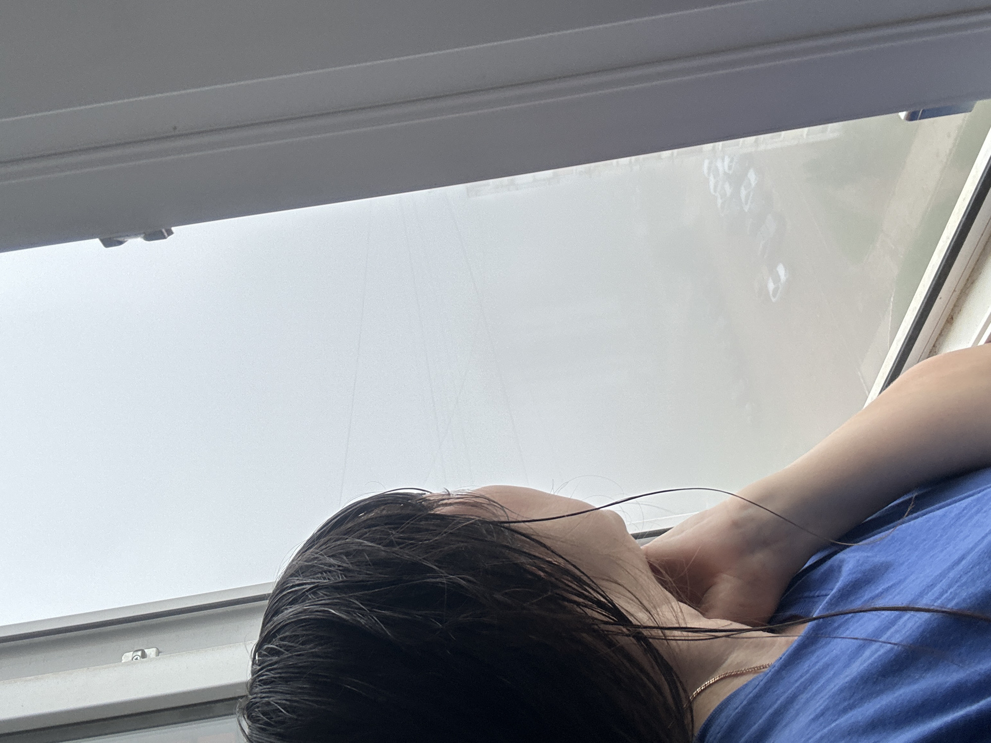 The image, taken in Chaikovsky, Russia, depicts a person lying on their side with their head tilted back, looking upwards towards a bright window or skylight. The primary subject is a person, likely female, seen from the top of their head to their upper torso. They have long, dark, wet-looking hair that falls across their neck and shoulders. They are wearing a blue t-shirt and a thin, golden-colored chain necklace is visible around their neck. Their right arm is bent, with their hand resting behind their head.
The setting appears to be indoors, next to or beneath a large window. The window frame, modern and gray, is prominent along the top and right edges of the image. Through the window, the outside view is extremely bright and appears overcast or hazy, with very faint, blurry shapes visible on the right side, possibly distant buildings or landscape features. The light suggests it is daytime. The activity of the person seems to be resting or looking contemplatively out of the window. There is no visible text in the image.