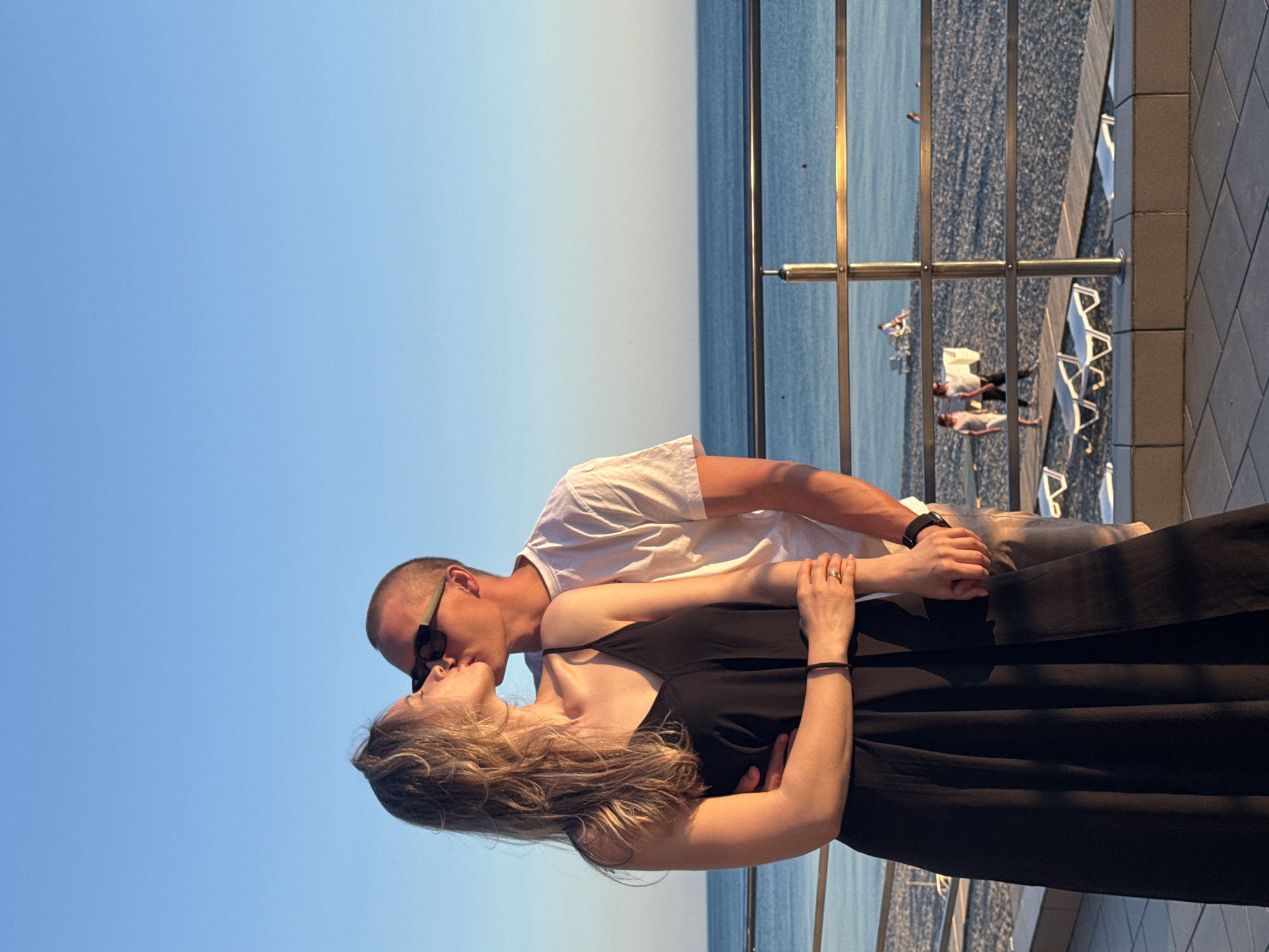 The image depicts a couple kissing on a promenade overlooking a pebble beach and the Black Sea in Nebug, Russia, during the golden hour. The woman, with blonde wavy hair, is on the left, wearing a black maxi dress. Her left hand rests on the man's arm, and her right hand holds his left, revealing a thin black bracelet on her left wrist and a ring on her left ring finger. The man, on the right, has short hair and wears a white t-shirt, light shorts, and dark sunglasses. He embraces the woman with his right arm around her back. A dark watch is visible on his left wrist. Both are leaning in for a kiss, their eyes closed.
Behind the couple, a silver metal railing separates the promenade from the beach. The beach is composed of small gray pebbles, with several white lounge chairs and a wooden walkway visible. In the distance, other individuals are present on the beach, some walking, others near lounge chairs. Beyond the beach, the calm, blue-green sea extends to the horizon, with faint red buoys visible on the water's surface. The sky above is a clear, bright blue, transitioning to a lighter hue near the horizon. The lighting is warm, casting soft light and mild shadows, indicative of late afternoon or early evening.