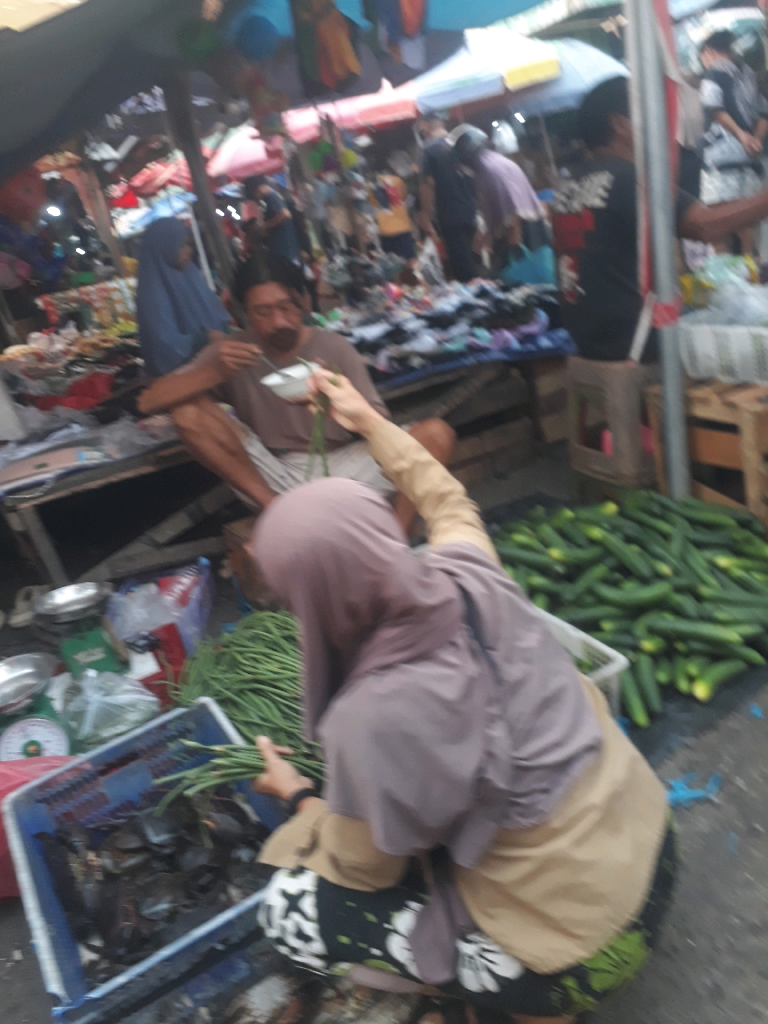 The image depicts a bustling street market in Pangkalpinang, Indonesia. In the foreground, a person wearing a light brown hijab and a tan overshirt is visible from the back, holding a bunch of green beans over a blue plastic crate. The crate contains what appear to be seafood items, possibly crabs or fish. To the right of the crate, a large pile of cucumbers is spread on the ground. Behind this individual, a man with a mustache, wearing a brown t-shirt and light-colored pants, is seated and appears to be eating from a white bowl with a spoon. He is looking down at his food. Further in the background, a woman in a blue hijab is visible, and other market stalls with various goods and people are blurred due to the image's focus and resolution. The overall environment suggests a lively outdoor marketplace with a variety of produce and goods being sold.