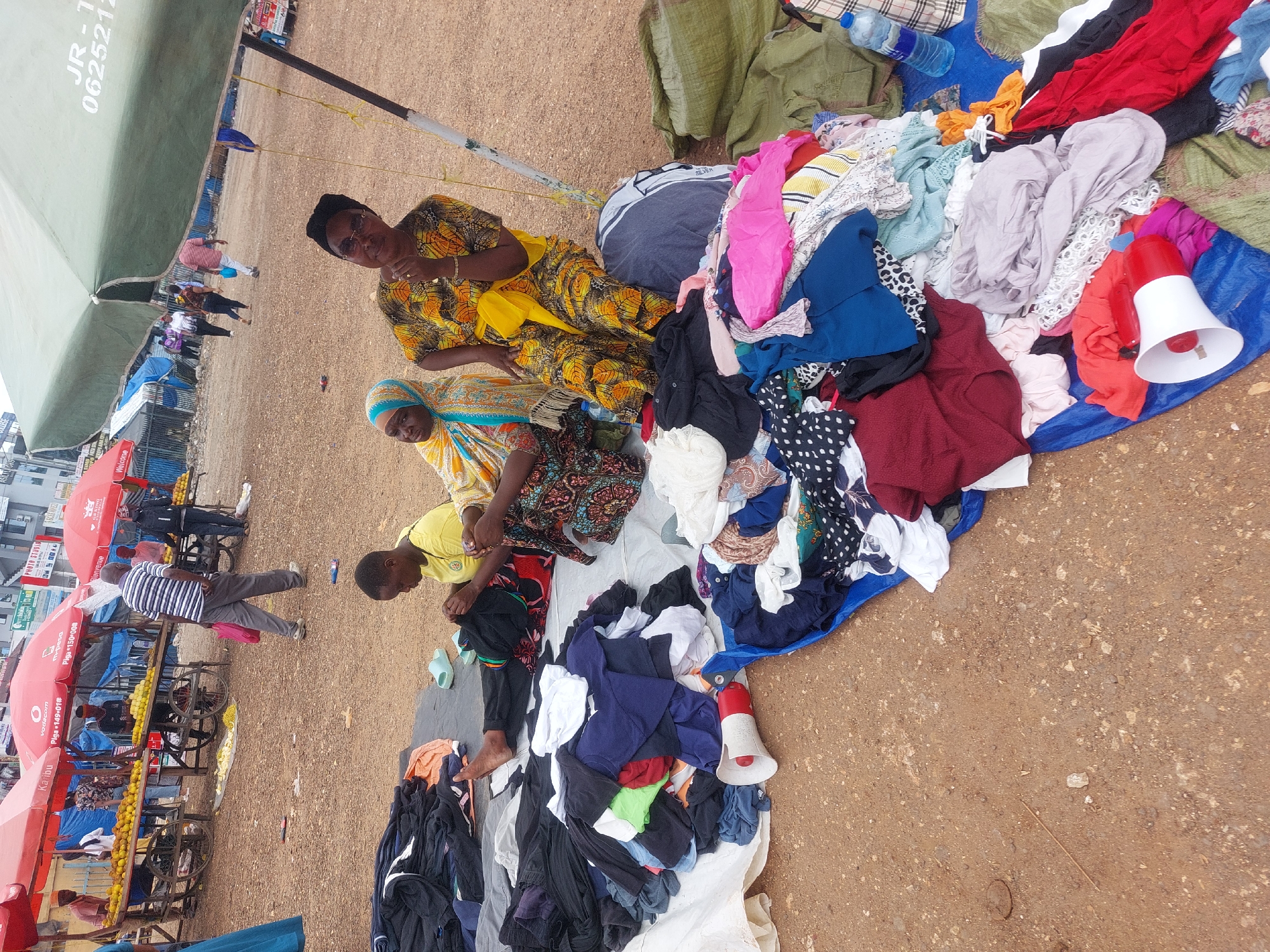 The image captures a bustling scene at a street market in Dar es-Salaam, Tanzania. Three women are seen attending to piles of clothing spread out on tarpaulins. Two are seated, one in a vibrant yellow and brown patterned dress, and another in a blue and yellow headscarf. A third woman, wearing a yellow shirt, appears to be sorting through the garments. The clothing ranges in color and style, from pink tops to polka dot dresses. A red and white megaphone rests among the clothing, possibly used to attract customers.
In the background, a row of vendors selling fruits and vegetables are set up under red umbrellas bearing the logos of Vodacom and M-Pesa. A man in a striped shirt walks through the market. Across the street, a building with a sign advertising a photo studio is visible. The ground is bare and rocky. A large green awning provides shade over the clothing vendors.