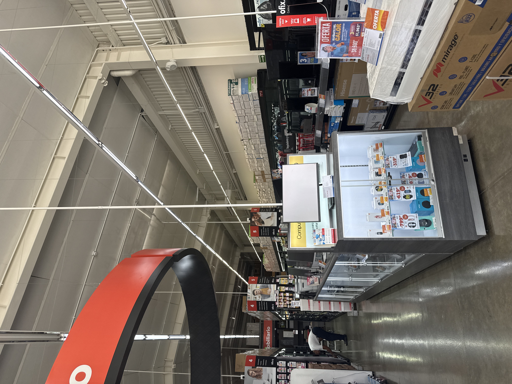 The image shows the interior of a large retail store, likely an electronics, appliance, or office supply outlet, located in San Bernardino Tlaxcalancingo, Mexico. The perspective is tilted, showcasing a high ceiling with exposed metal structures and bright linear fluorescent lights.
In the foreground, a prominent red and black arch-like structure frames the left side of the view. The store features multiple product displays, including a long glass display case filled with small electronics and accessories like smartwatches and "echo pop" devices, marked with prices such as "$799", "$619", "$819", and "$319". Further into the store, shelves are stocked with various items, some bearing the "Ofix" brand.
Large appliance boxes, specifically "Mirage" and "Hisense" air conditioning units, are displayed. A significant "OFERTA" (Offer) sign advertises a "MINISPLIT INVERTER" unit with the slogan "OLVÍDATE DEL CALOR" (Forget the heat) for "$6,690" down from "$5,890.00", and a "3 MESES" (3 months) payment option. Another sign indicates "COMPRA AQUÍ" (Buy here). A person, potentially a shopper or employee, is visible bending down near a lower shelf in the background. Overhead, a security camera is mounted.
Visible text includes brand names like "Ofix," "Hisense," "Mirage," and "echo pop." Promotional text includes "OFERTA," "UHD 4K," "MINISPLIT INVERTER," "R32," "INVERTER," "OZONE CARE," "LIBRARY APP," "PRO-PLANET," "X-COOL," and prices. Section signs like "mobiliario" (furniture), "Reguladores y," and "Compu para soñar" (Computer for dreaming) are also present, along with numbered aisle markers 4, 5, and 6. The store has polished concrete flooring and industrial-style lighting, suggesting a spacious, modern retail setting.