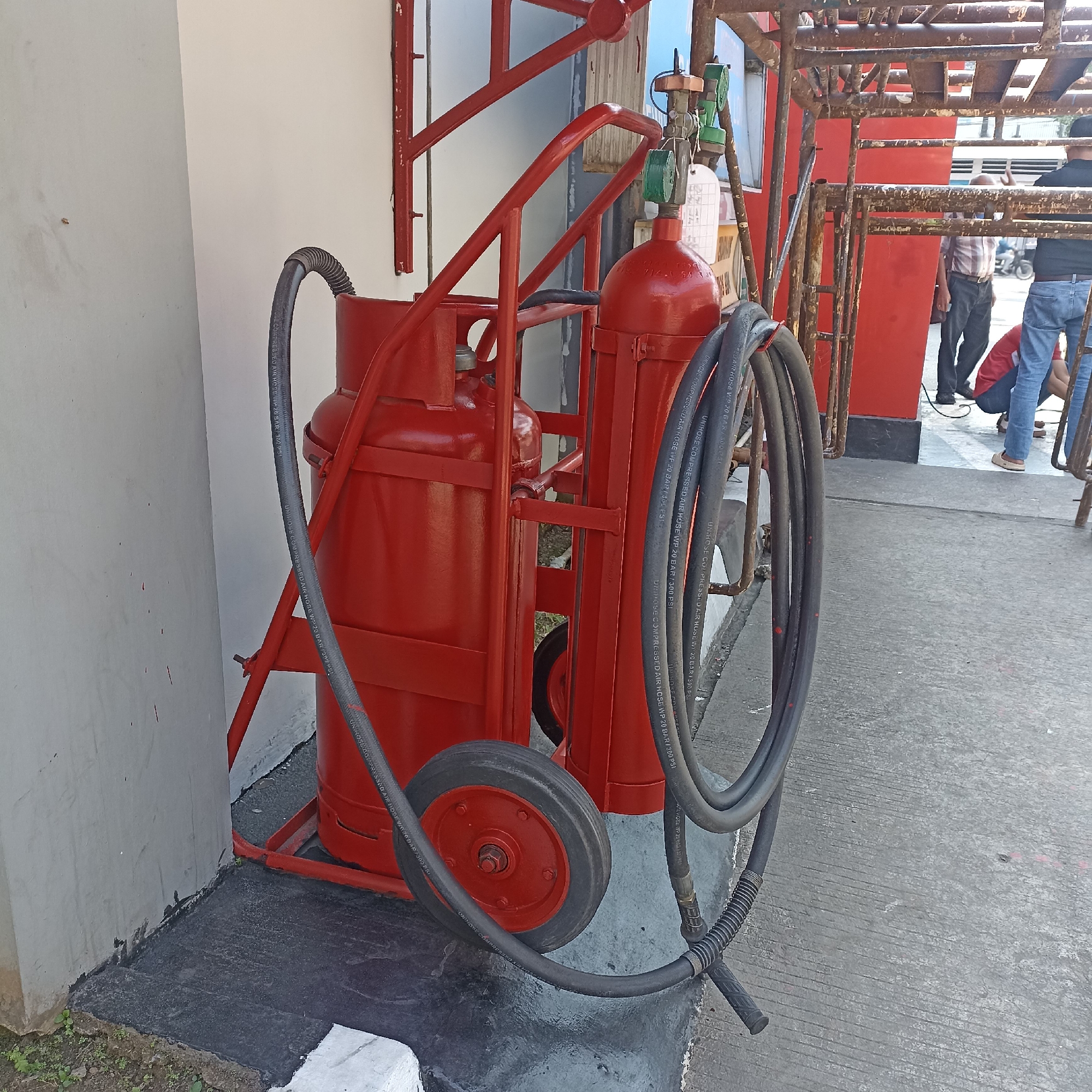 A red wheeled cart with two red tanks and a coiled black hose is parked next to a white wall. The cart has a red metal frame. The hose is coiled around the tanks. In the background, people are visible near a red metal structure. The scene appears to be outdoors on a concrete surface. This image was taken in Magelang, Indonesia.