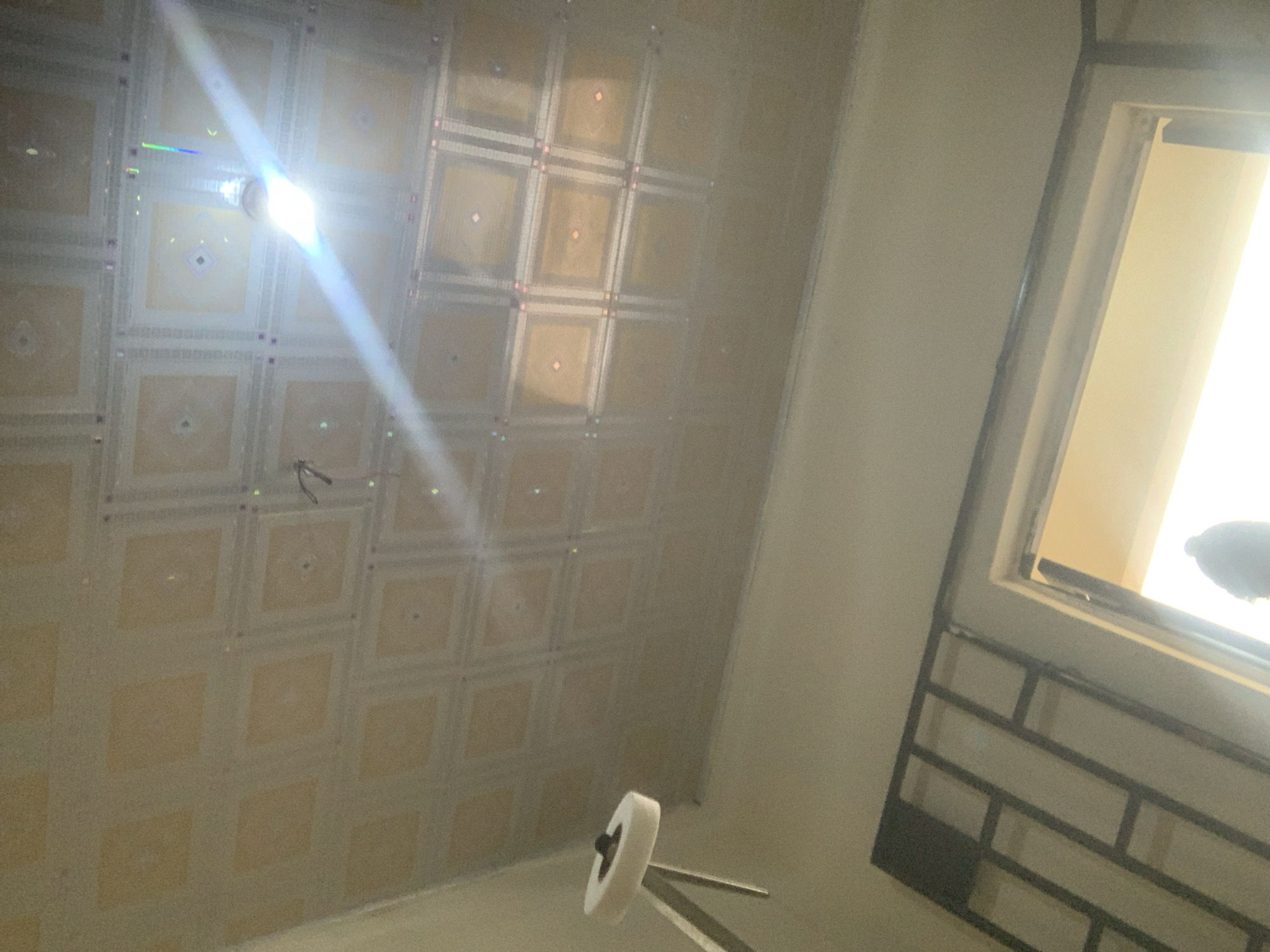 The image shows an interior view of a room with a patterned ceiling. A light fixture is visible on the ceiling, emitting a bright light and a lens flare. A white object with a circular base and a smaller dark object on top is positioned on the left side of the image, possibly a lamp or part of a lighting fixture. The lower part of the image reveals a window with natural light streaming in. A dark metal grate or bars are visible against the window. A silhouetted figure is partially visible in the window opening. The walls are a neutral color. The overall impression is of an indoor space, possibly under construction or renovation, given the exposed elements and lighting. There is no specific indication of the city of Jauro Yinu, Nigeria within the image itself.