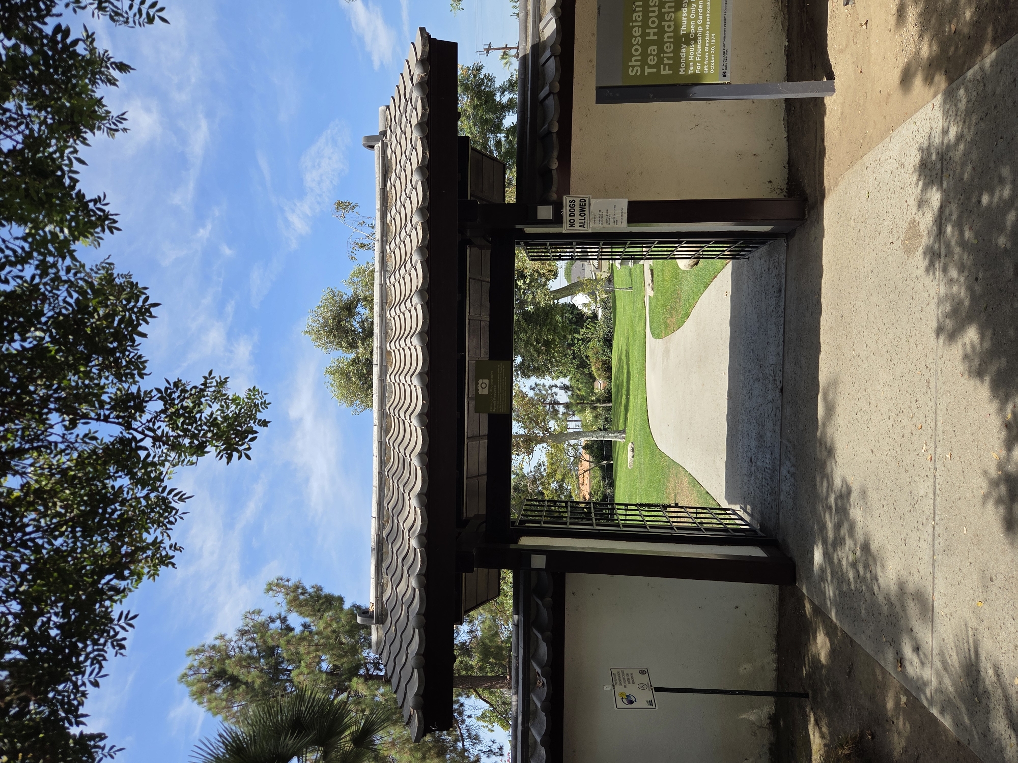 The image shows the entrance to the Shoseian Tea House and Friendship Garden in Glendale, California. The entrance features a traditional Japanese-style gate with a tiled roof and wooden beams. The gate is open, revealing a paved walkway leading into a lush green garden.
On the left side of the entrance, there is a white wall with a sign that reads "SUPPORT CONSERVATION WE USE WATER FOR IRRIGATION". There is also a circular emblem with a camera icon and a line through it, indicating that photography is not allowed or requires a permit.
On the right side of the entrance, there is a sign with the text "NO DOGS ALLOWED". Below this, another sign reads "Shoseian Tea House Friendship Garden". The opening hours are listed as "Monday - Thursday" and "Tea House Open Only", with a note that it is a "gift from Glendale Higashiosaka Sister City Association October 20, 1974". A small camera icon and a phone number are also present.
The garden itself is well-maintained, with green grass, trees, and a winding pathway. The sky is blue with scattered white clouds, and sunlight filters through the leaves of overhead trees, casting dappled shadows on the ground. The overall scene conveys a sense of tranquility and cultural heritage.