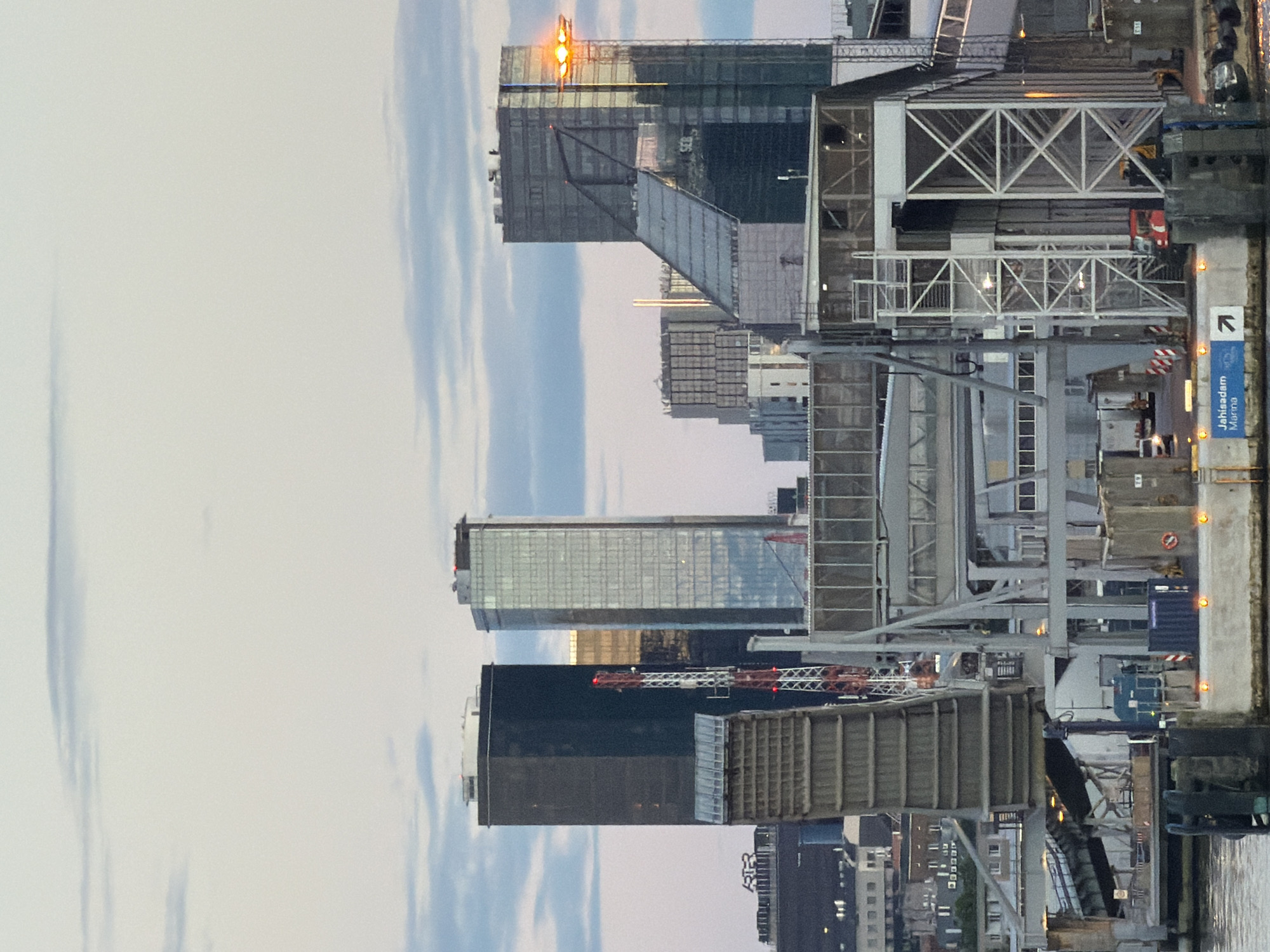 The image depicts a harbor in Tallinn, Estonia, with a focus on the ferry terminal area and the city's modern skyline. In the foreground, a raised drawbridge or ramp for vehicles is visible, with concrete bollards and various industrial equipment, including containers and what appears to be a blue truck or cargo vehicle. Several orange lights are intermittently placed along the pier. A blue shipping container with white lettering is also present. In the background, a cluster of contemporary skyscrapers dominates the horizon, including a tall, dark rectangular building and a taller, glass-covered building with a distinctive angular design. A red and white communication tower stands between these two main buildings. The sky above is partly cloudy, with soft, diffused light suggesting either early morning or late evening. The scene is devoid of any discernible human figures.