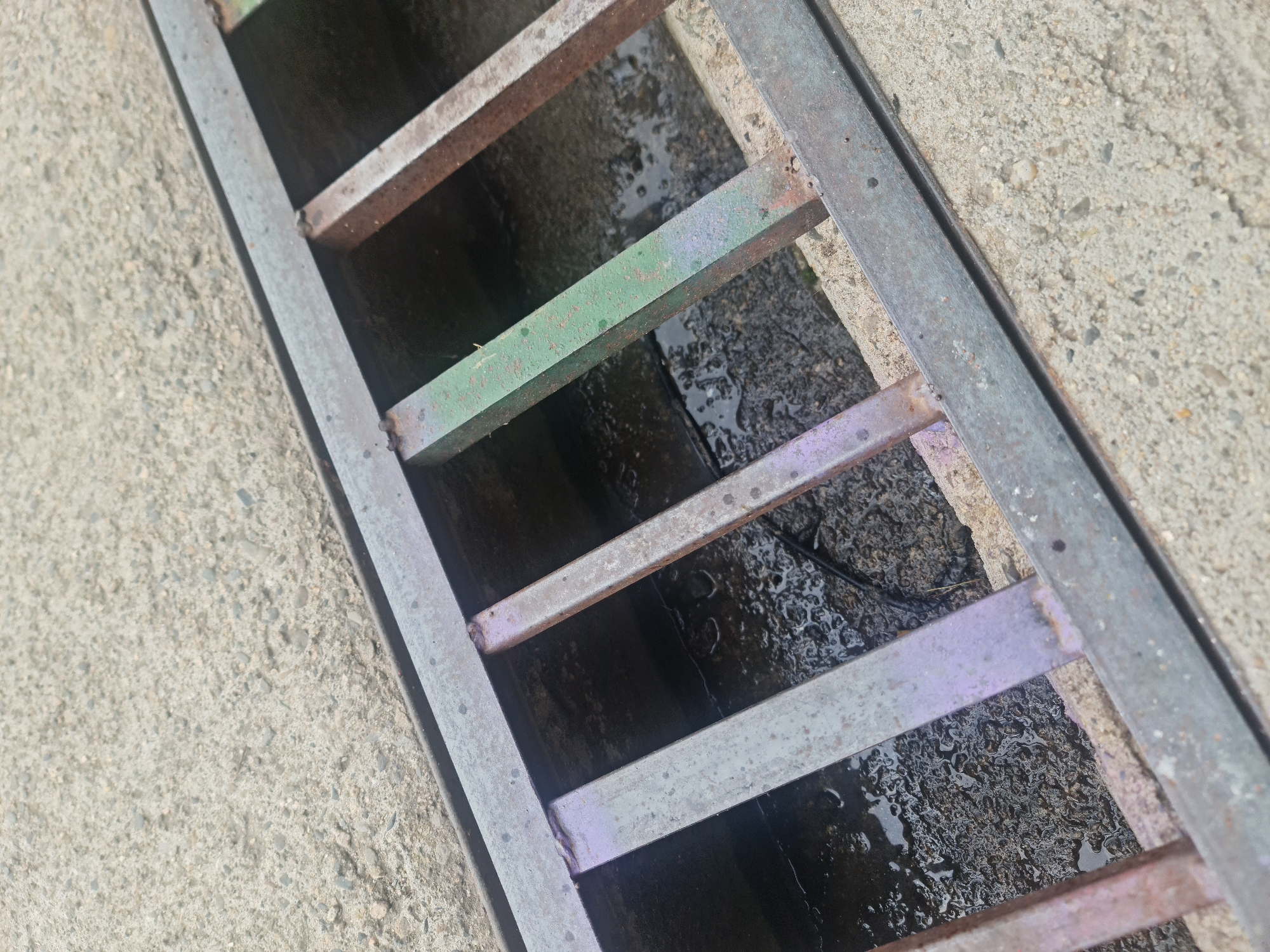 This close-up, angled image captures a weathered metal grate covering a drainage channel or trench. The grate consists of several parallel metal bars, some of which bear remnants of faded paint in shades of green, purple, and reddish-brown, suggesting rust or old paint. These bars are welded onto two thicker, parallel metal supports that frame the opening. The metal components show signs of age and wear, including corrosion.
The grate is set into a light gray, granular concrete surface, which appears dry and has a rough texture with visible small pebbles. Below the grate, the drainage channel is dark and wet, containing standing water that reflects light unevenly, creating shiny, rippled patches. The bottom of the channel appears to be composed of dark, possibly muddy or debris-filled ground. A thin, dark, possibly electrical, cable or wire is faintly visible within the channel in the upper right section.
The lighting indicates daytime, with a clear, ambient illumination. There are no people present, nor is any specific activity taking place. The scene depicts a static piece of infrastructure in an outdoor setting, consistent with an urban or semi-urban environment in Buzovgrad, Bulgaria. No visible text is present in the image.