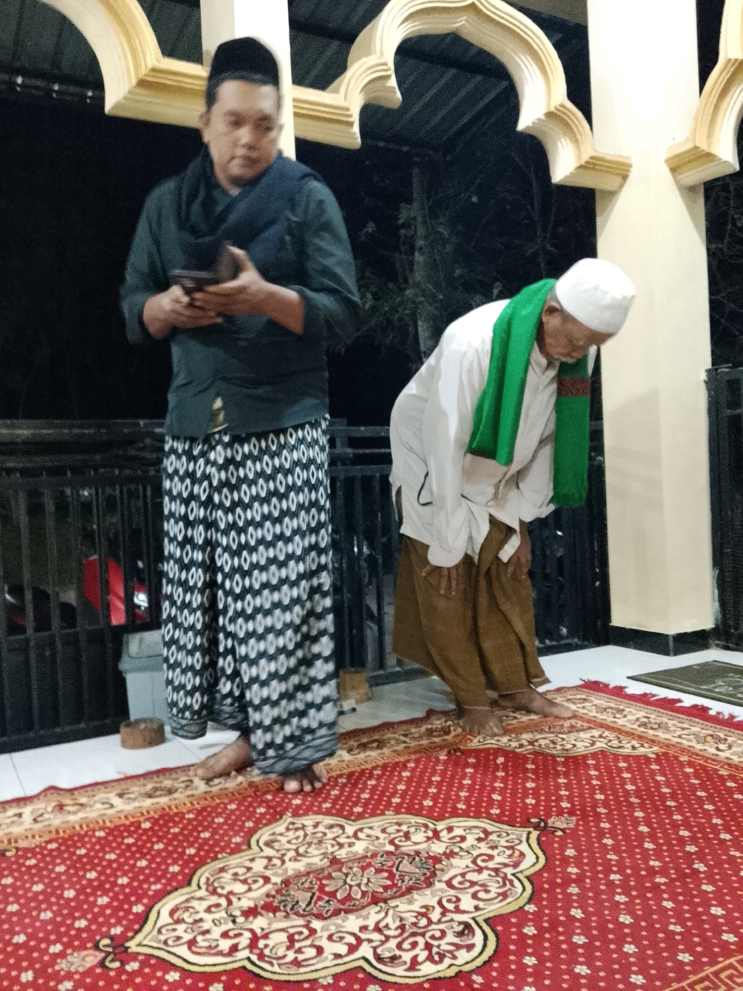 Two men are standing on a red carpet with an ornate pattern. The man on the left, wearing a dark green shirt, a dark blue scarf, and a black peci (cap), is holding a dark-colored smartphone in his hands. He is also wearing a black and white patterned sarong that extends to his ankles. His feet are bare. The man on the right, dressed in a white shirt, a green scarf, and brown trousers, is in a bowing position, with his hands on his knees. He is wearing a white peci (cap). A second, smaller, darker mat is visible in the background to the right of the red carpet. The scene appears to be outdoors, possibly in a courtyard or veranda, with decorative architectural elements, including curved arches, visible in the background. The lighting suggests it is nighttime.