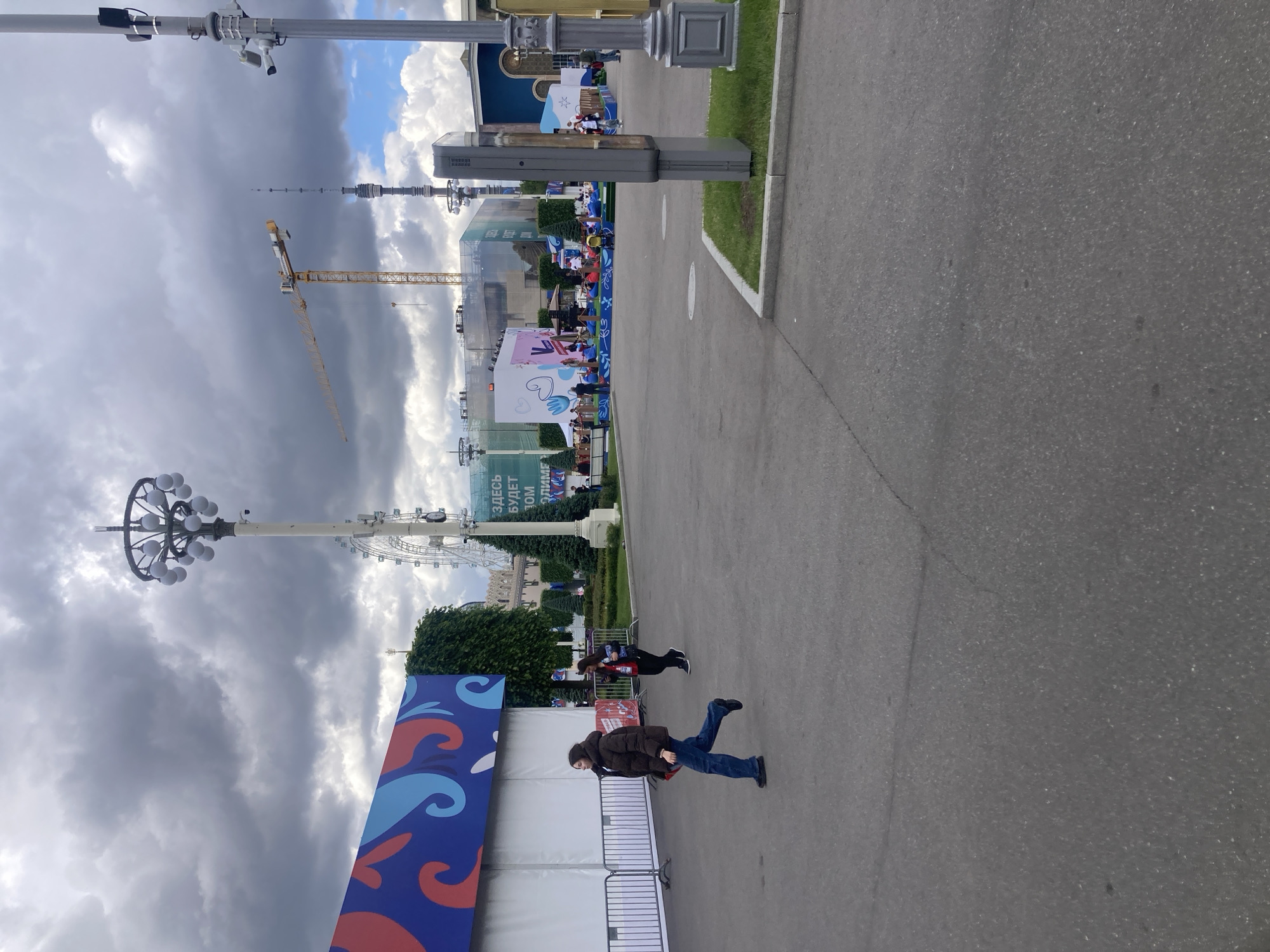 The image depicts a public space in Moscow, Russia, near the Ferris wheel and Ostankino Tower. The foreground shows a paved pathway. On the left, there is a large banner with a blue and red design and a white fence. Two people are walking along the path. A person in a brown jacket and blue jeans is closer to the camera. Another person, wearing a black jacket and pants, is further away. Overhead, the sky is cloudy with a mix of dark and bright clouds. Visible in the background are the Ferris wheel, a construction crane, and the Ostankino Tower. There are signs with Russian text visible along the path.