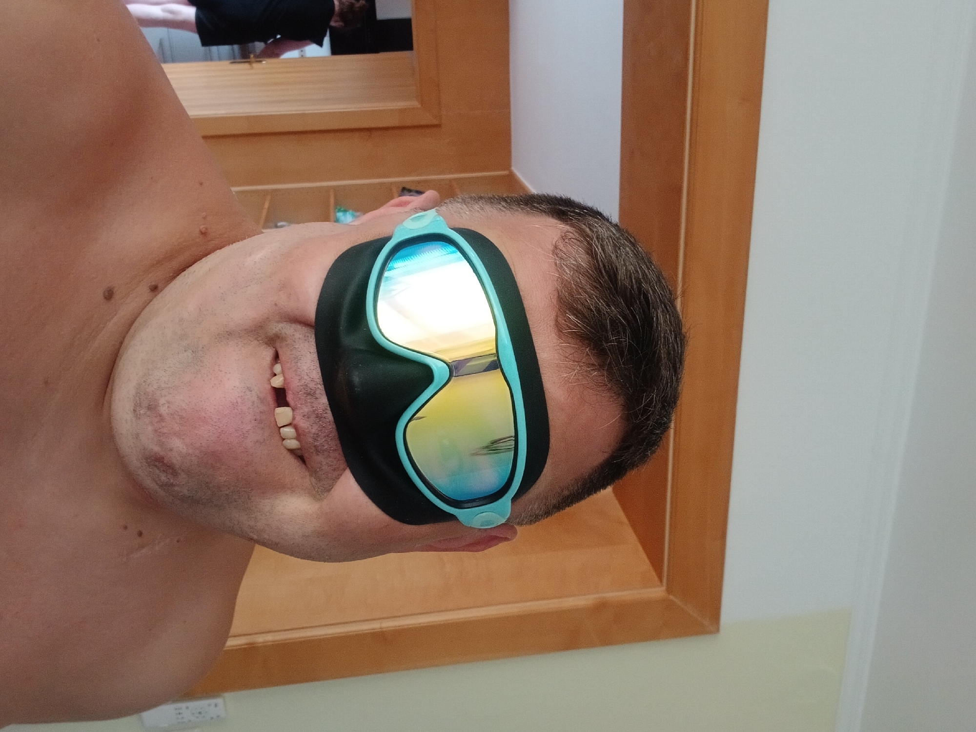 The image shows a close-up of a person wearing a pair of swimming goggles, obscuring their eyes. The goggles are light blue and have mirrored lenses reflecting yellow and blue colors. A black facial mask is worn underneath the goggles, covering the nose and area around the eyes. The person has short, dark hair, and their mouth is visible, revealing missing teeth as they smile. The background appears to be an indoor setting with wooden-paneled doorways and walls. In the far background, a person in a black dress is partially visible walking through a doorway. There are shelves visible in the background as well. The image does not contain any information about the location of Pfarrkirchen, Germany.