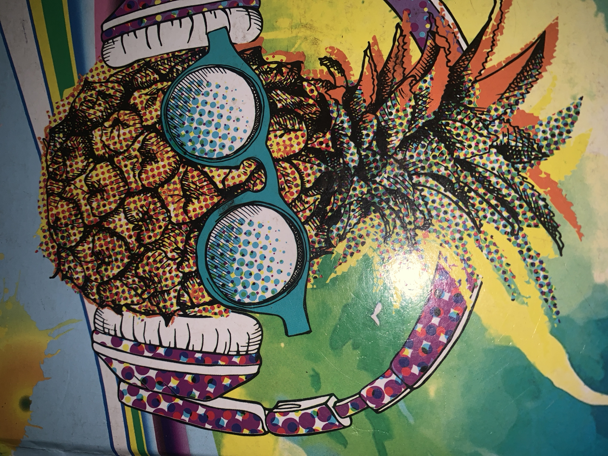 This is a photograph of a colorful illustration of a pineapple wearing headphones and sunglasses. The pineapple is depicted in a pop art style, with a dot pattern filling its texture and the leaves. The headphones have a pattern of purple, pink, and white circles. The sunglasses are teal, with a dotted pattern inside the lenses. The background is a gradient of green and yellow, with some abstract shapes and lines in blue, yellow, green, and pink. The image appears to be a piece of artwork, possibly a poster or a print. There are no people or specific city locations depicted in the image.