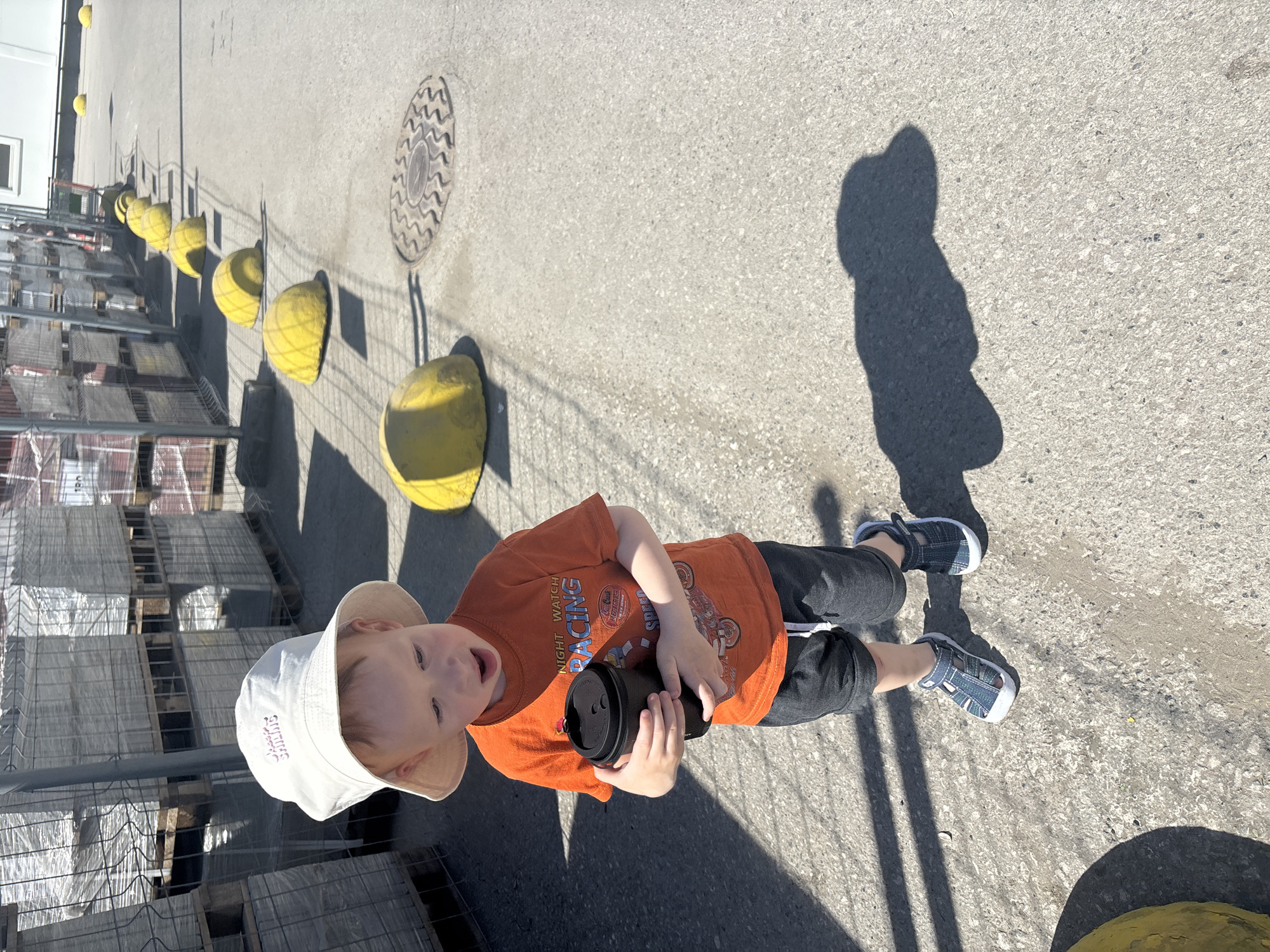 A high-angle shot depicts a toddler holding a black tumbler standing on a concrete surface. The child is wearing a white bucket hat, an orange t-shirt with graphics, dark gray pants, and closed-toe sandals. The toddler is positioned slightly off-center, facing the camera.
To the left of the child is a stacked pallet of materials, possibly bricks or concrete blocks, encased in wire caging. A row of yellow bollards, evenly spaced, stretches out along the left side of the frame, casting shadows on the concrete. A manhole cover with a circular pattern is visible in the background. The toddler casts a shadow on the concrete to the right.
The location is an outdoor paved area, likely a construction supply or hardware store, in what could be Perm, Russia. The buildings and objects are of a utilitarian construction style common in many urban industrial areas. The light suggests a sunny day.