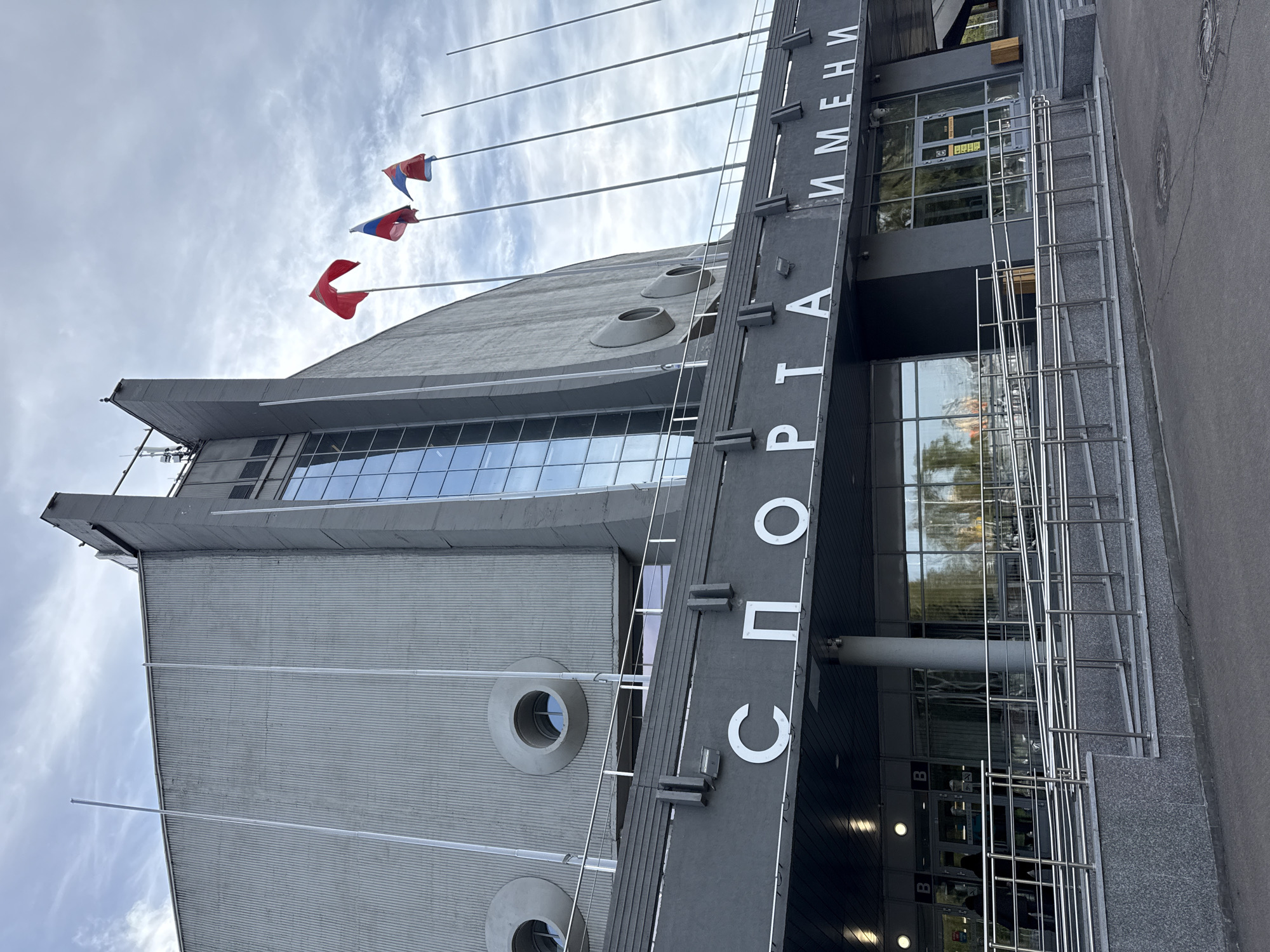 The image shows the front of the Ivan Yarygin Sports Palace in Krasnoyarsk, Russia. The building is a modern architectural structure with a grey facade, large windows, and circular recessed elements. A sign with white Cyrillic lettering spelling out "ДВОРЕЦ СПОРТА ИМЕНИ" (Palace of Sports named after) is prominently displayed. Several flagpoles extend from the building, with red, white, and blue flags, possibly Russian Federation flags, fluttering in the sky. A metal railing leads towards the entrance, where glass doors and windows reflect the surroundings. The scene is captured from a low angle, looking upwards towards the building and sky. The sky is overcast with scattered clouds. A portion of the paved ground is visible in the foreground.