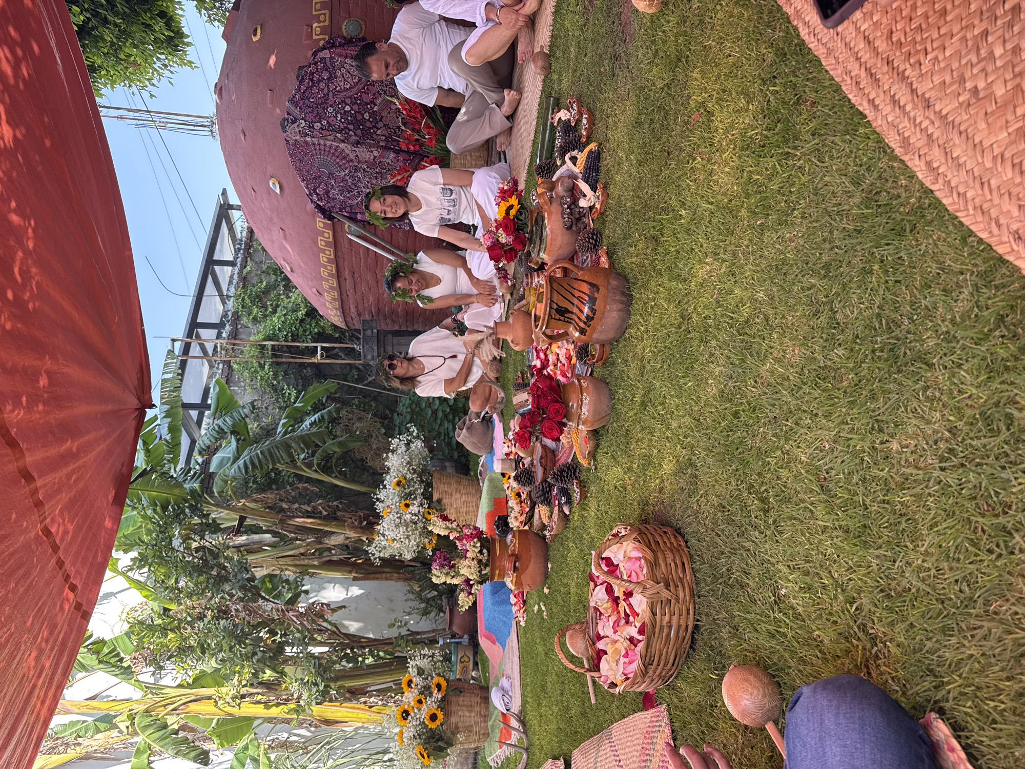People are sitting outside in a yard in Puebla, Mexico. Four people are seated on the ground, wearing white shirts. One man is sitting behind them, wearing a white shirt and brown pants. The people on the ground have flower crowns on their heads. There are baskets of flowers and pottery jars of flowers and food in front of them. A large red-colored dome structure is behind them.