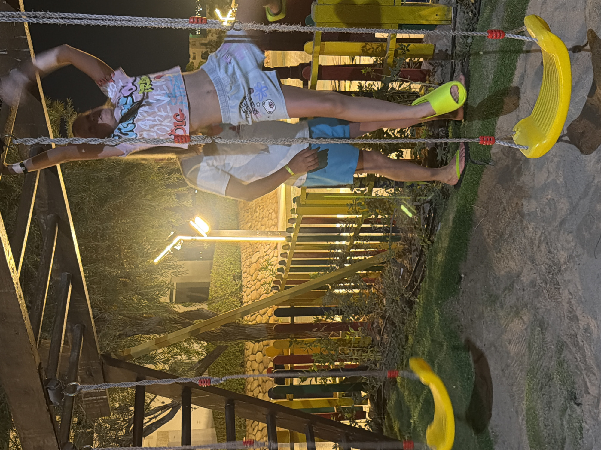 In Sharm El Sheikh, Egypt, a young girl with blonde hair is suspended from a playground structure by ropes, her arms outstretched as she grips the top bar. She is wearing a colorful graphic t-shirt and light-colored shorts. Below her, a person wearing a white t-shirt and blue shorts stands on the grass. The playground features yellow swings. The area is lit by artificial lights, casting a warm glow on the scene and creating shadows. The background includes a low, multi-colored wooden fence and a stone wall. The ground is a mix of sand and grass.
