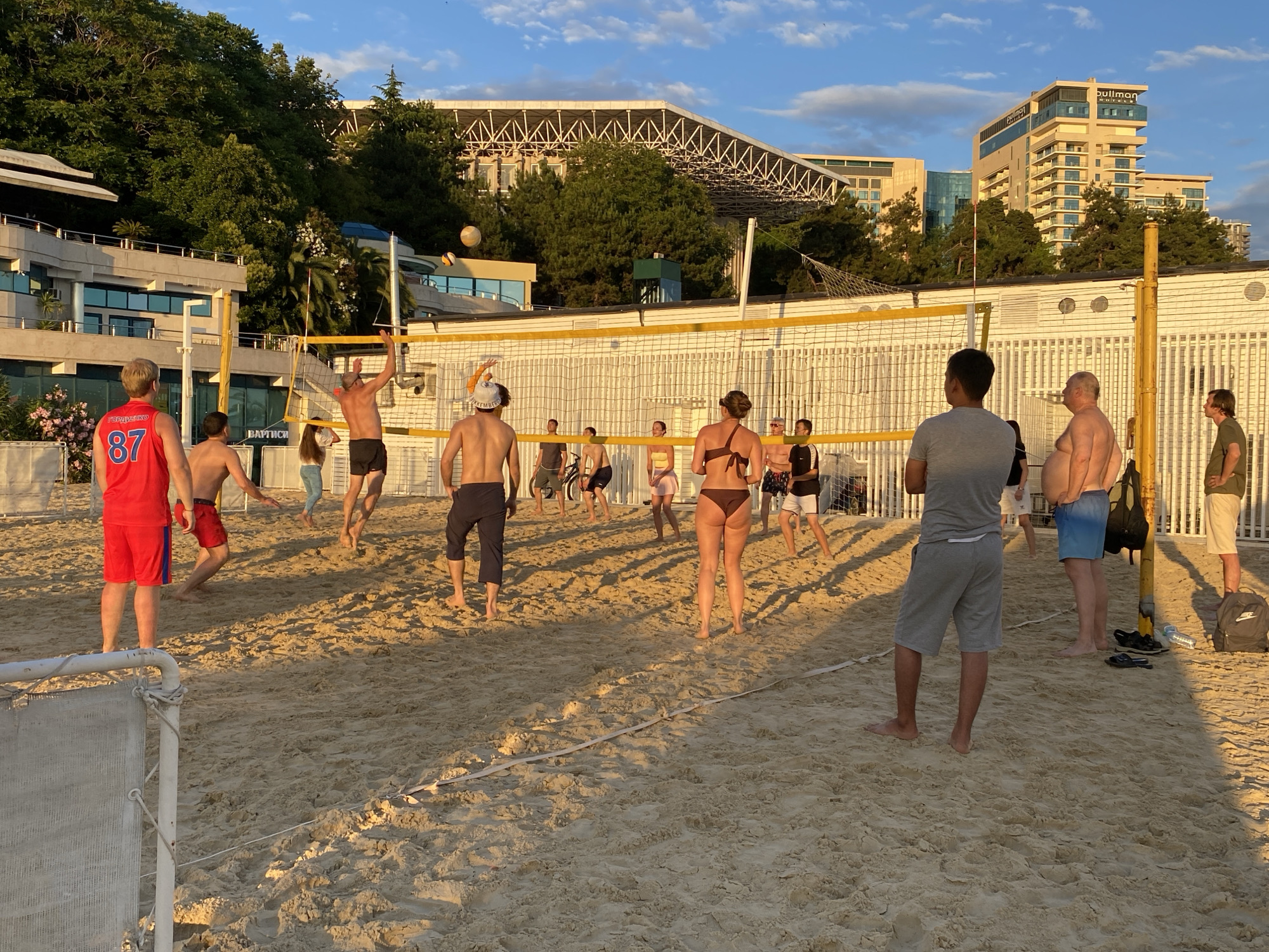 The image shows a group of people playing beach volleyball on a sandy court. The sun is setting, casting long shadows across the sand, indicating it is late afternoon or early evening. The sky is a clear blue with a few scattered clouds. In the background, there are large modern buildings, possibly hotels or resorts, nestled among lush green trees. A distinctive white structure with a metal frame, resembling a stadium roof, is visible on top of a hill.
The people are dressed in beach attire. Several men are wearing shorts, and some are shirtless. One man on the left is wearing a red tank top with the number "87" on it. Some women are in bikinis. They appear to be actively engaged in the game, with some jumping to hit the ball and others positioned on the court. The volleyball net is stretched across the sand, and the net poles are visible. A volleyball is in mid-air, suggesting the game is in progress.
On the left side of the image, there's a glimpse of a white structure that might be part of a beach club or facility, with some seating. A white mesh barrier is also visible in the foreground on the left.
The overall mood seems relaxed and recreational, characteristic of a summer evening activity at a beach resort. Some backpacks and a water bottle are on the sand near the net poles, indicating the players have brought their belongings. There is no visible text that is clear enough to read. The scene appears to be in Sochi, Russia, given the architectural style and the likely resort setting.