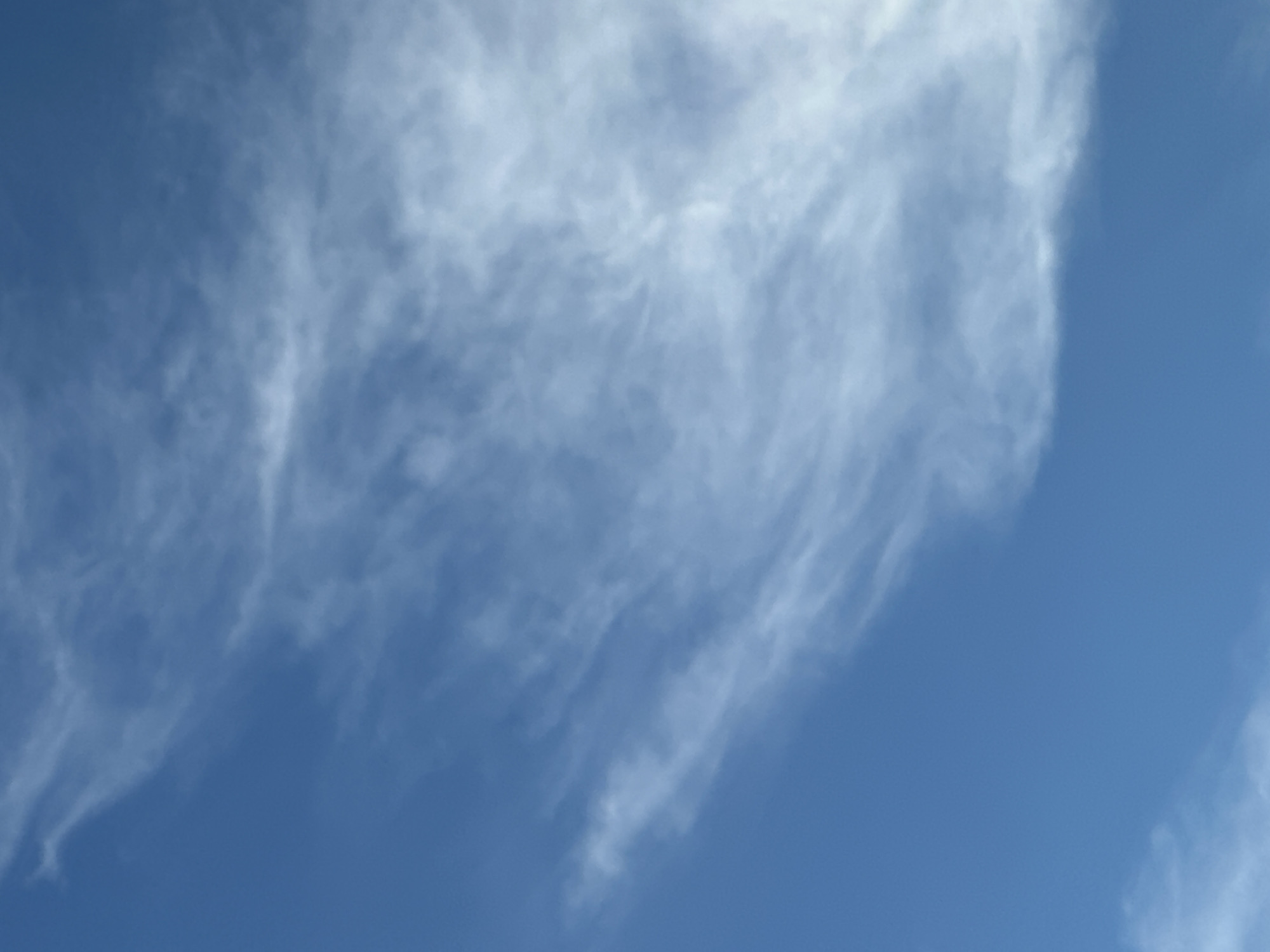 This is an image of the sky with scattered clouds. The sky is a vibrant blue. Thin, wispy white clouds streak across the frame, creating a textured effect. Some clouds are more dense and opaque, while others are translucent. The clouds appear to be moving, suggesting a windy day. There are no other objects or subjects visible in the image, such as buildings, people, or animals. The image is taken from a low angle, looking upwards at the sky. The lighting suggests it is daytime, with sunlight filtering through the clouds. The location is unknown.