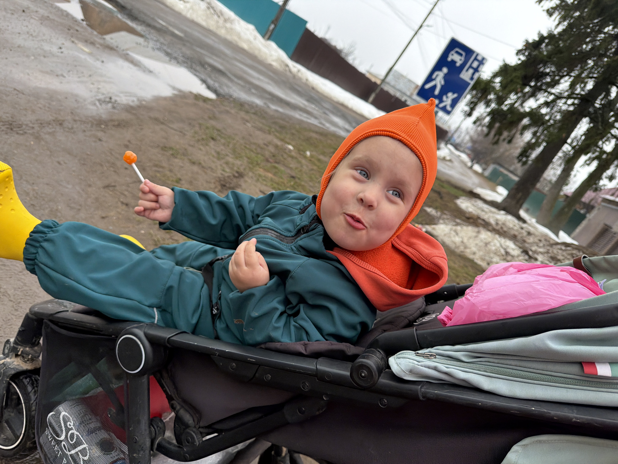 A child is sitting in a stroller outdoors. The child is wearing yellow boots, a teal jumpsuit, and an orange hat. The child is holding an orange lollipop in one hand. The stroller is black and contains a black basket below the seat. There is a bag with a pink plastic bag on top and a gray fabric bag with a red and white stripe. In the background, there is a road with snow on the side, a road sign, trees, and a fence. This photo was taken in Chaikovsky, Russia.