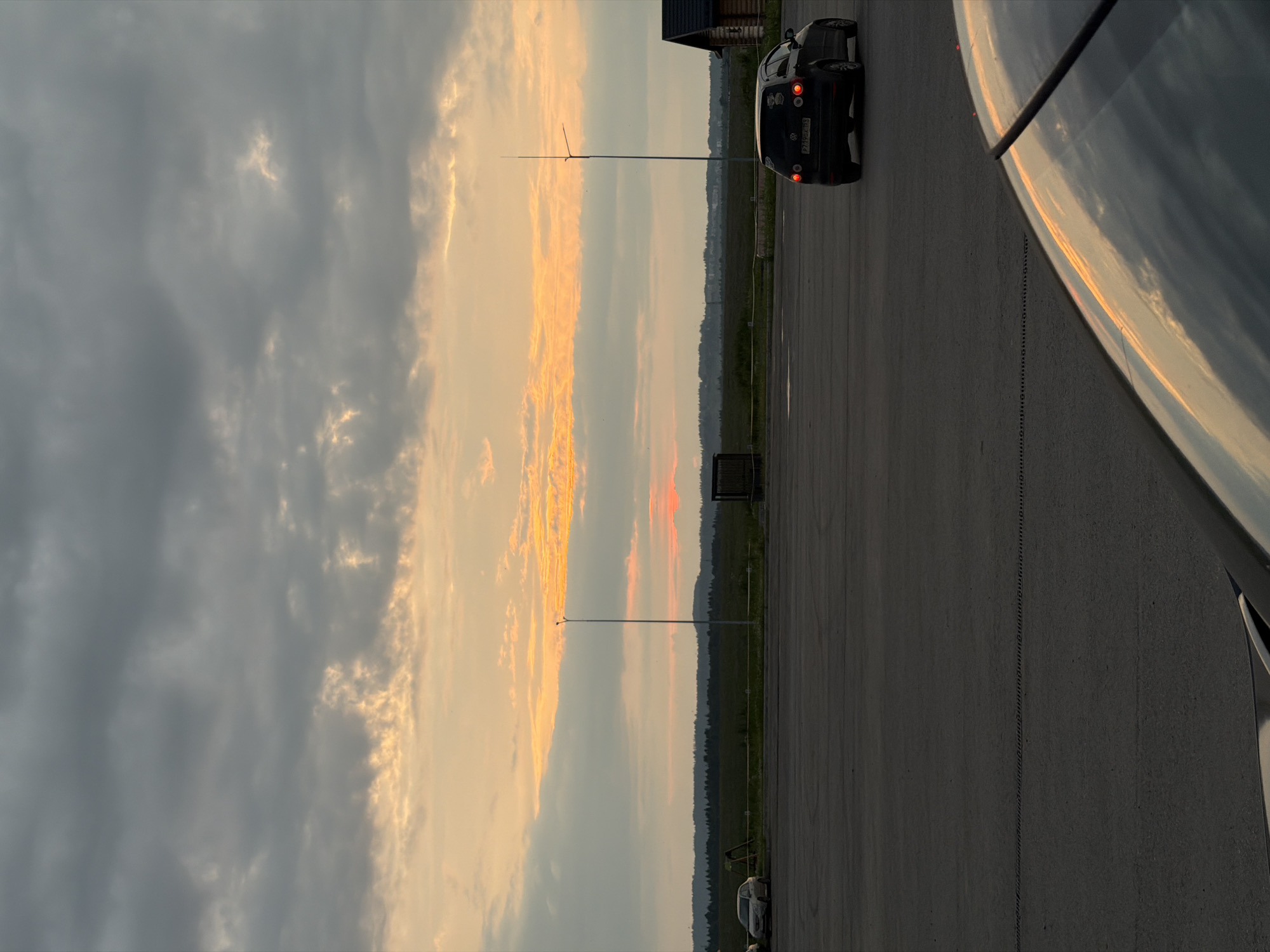 The image shows a sunset scene on what appears to be an airfield or an open, paved area in Ust-Sysolsk, Russia. A dark-colored car is parked on the right side of the frame, facing away from the viewer and towards the horizon. The sky is filled with dramatic clouds, with a band of bright orange and yellow light indicating the setting sun. The rest of the sky is a mix of dark gray and lighter, wispy clouds.
In the background, a thin, tall pole or antenna stands in the distance. Further beyond, a line of trees and hills is visible against the horizon, suggesting a rural or semi-rural setting. The ground is paved, with faint lines suggesting parking spaces or a road. The right side of the image is dominated by the reflection of the sky and clouds on a shiny surface, possibly the hood or roof of another vehicle.
The time of day is clearly sunset, indicated by the vibrant colors in the sky. The weather appears to be partly cloudy. There is no discernible human activity in the scene. The overall mood is peaceful and serene, capturing the beauty of the natural light at dusk.
Visible text on the car's license plate appears to be "P719HT 154".