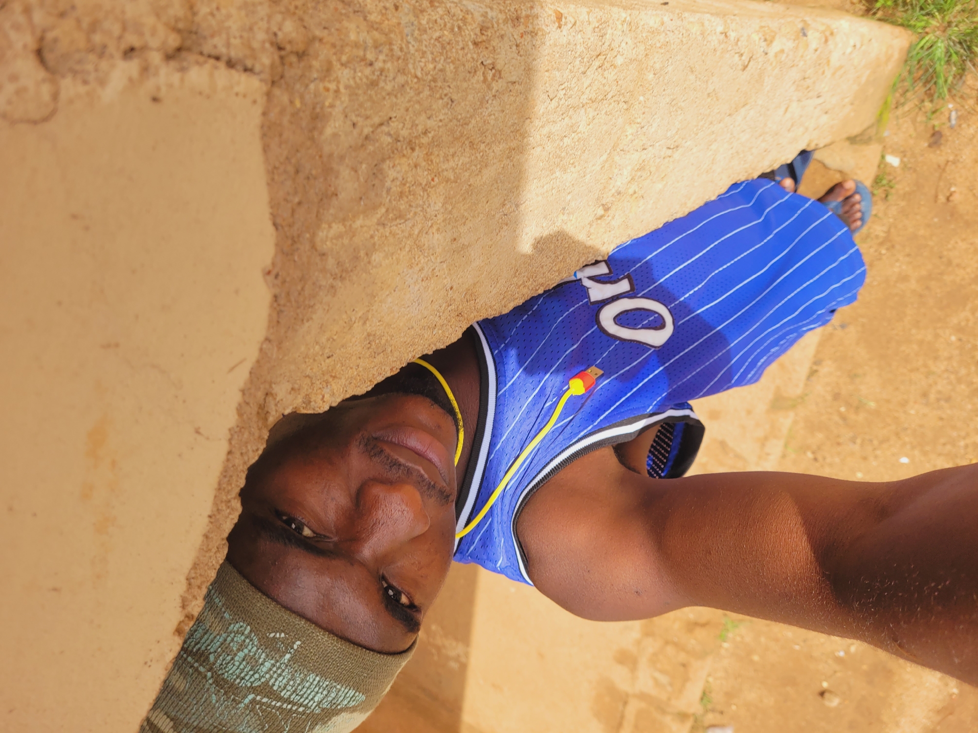 A selfie, taken from a low angle, shows a young Black male from the chest up, leaning out from behind a rough, tan-colored wall. He is wearing a green kufi hat and a blue basketball jersey with white pinstripes and the word "Or" visible on the front. A yellow cord, with a red and blue USB connector attached, hangs around his neck. His left arm is extended forward, holding the camera. The background is blurry, showing a sandy, dirt ground with some patches of green grass. The lighting suggests it is daytime, with sunlight casting a shadow on the wall. The environment appears to be outdoors, possibly in a rural or less developed area. The provided coordinates place this image in Bukuru, Nigeria.