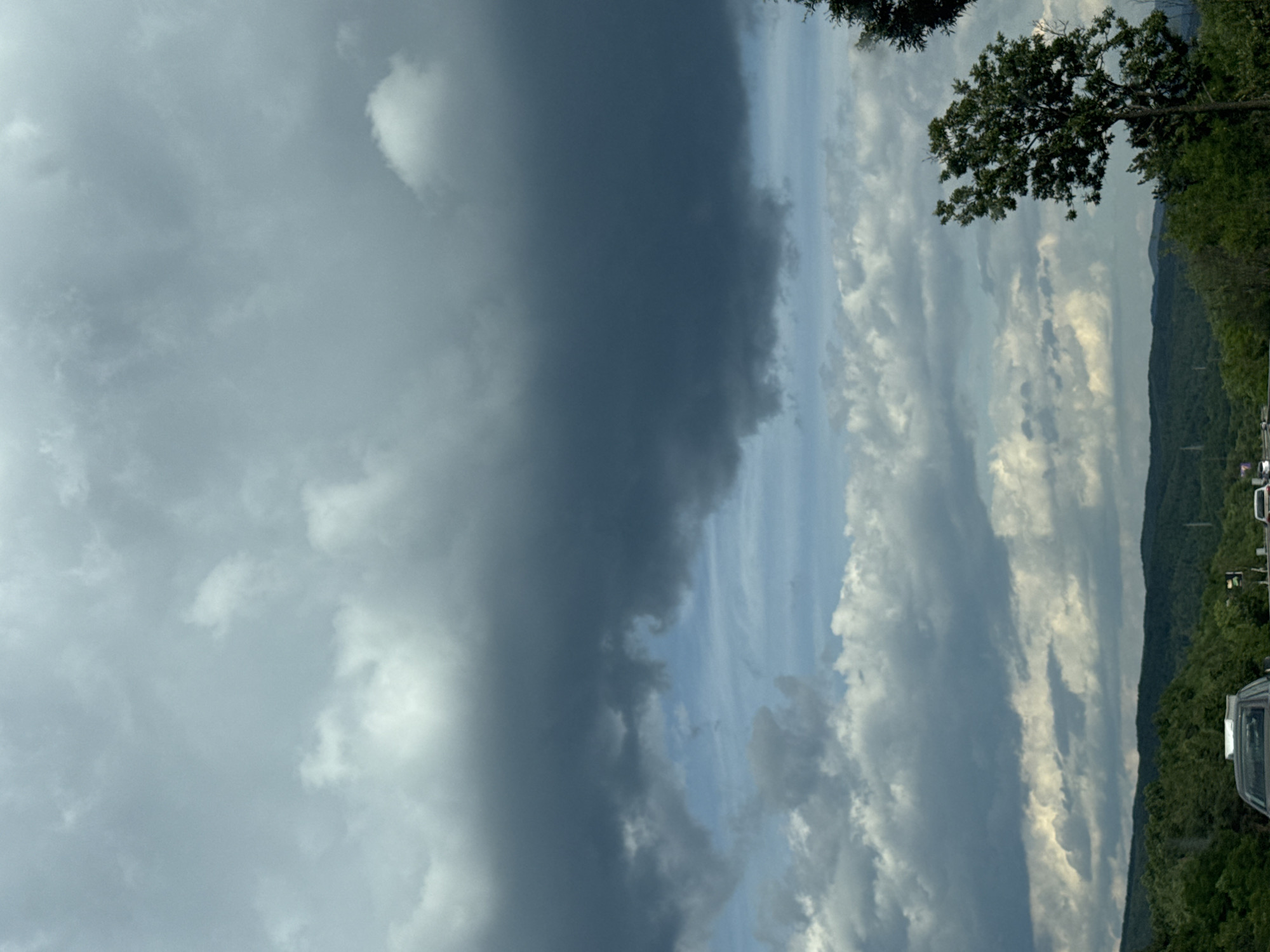 The image depicts a cloudy sky over a hilly, tree-covered landscape. In the foreground, a road with several cars is visible. The sky is filled with a variety of clouds, ranging from light and wispy to dark and heavy. The dark clouds suggest the possibility of rain or a storm. The hills are covered in dense green trees, and the overall impression is of a natural, rural setting. The location is not specified in the image.