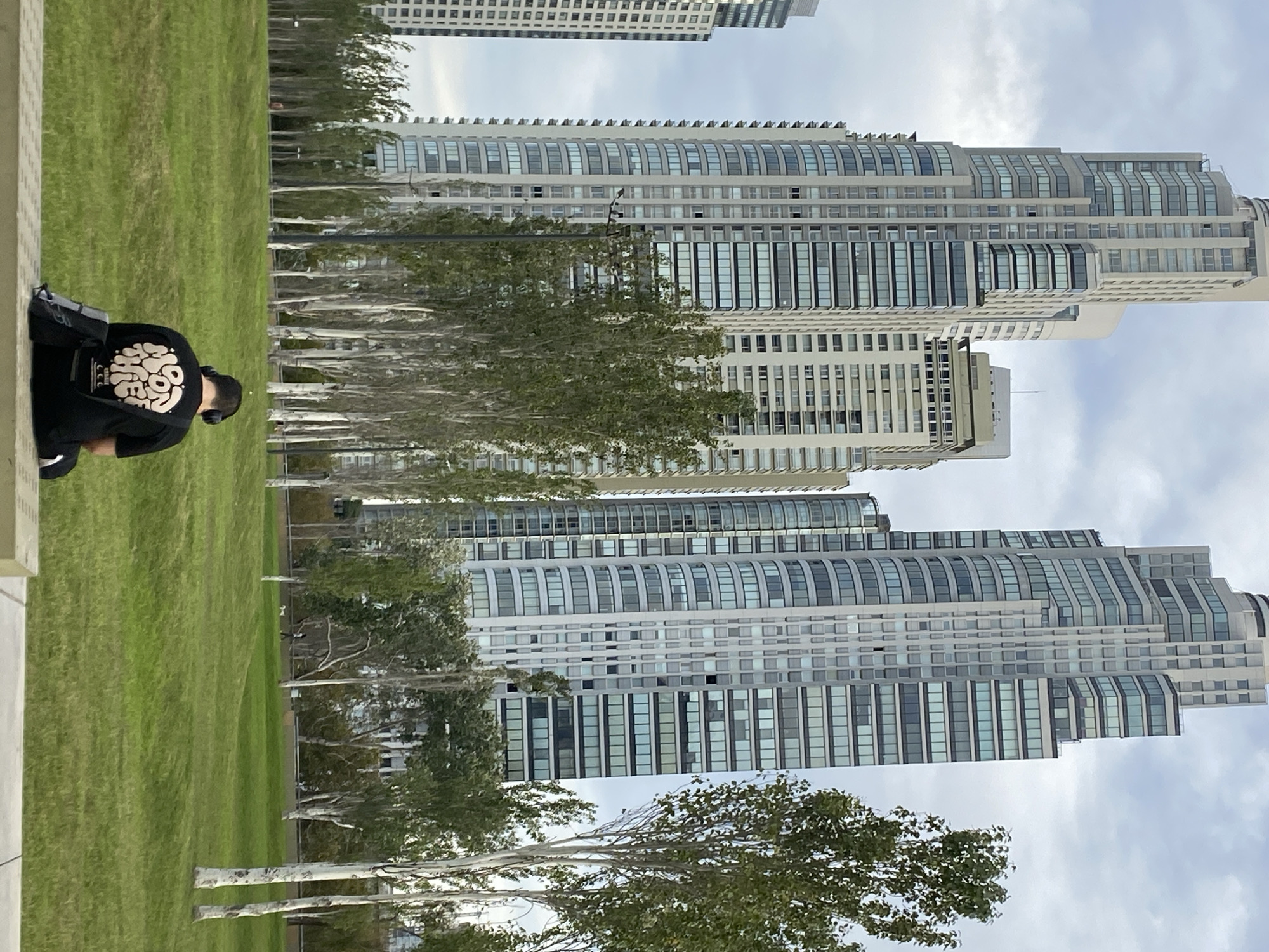 A man wearing a black shirt and headphones is sitting on a concrete ledge. Behind him is a grassy area with several birch trees. In the background are high-rise buildings. The location is likely in Buenos Aires, Argentina, given the architecture and urban landscape.