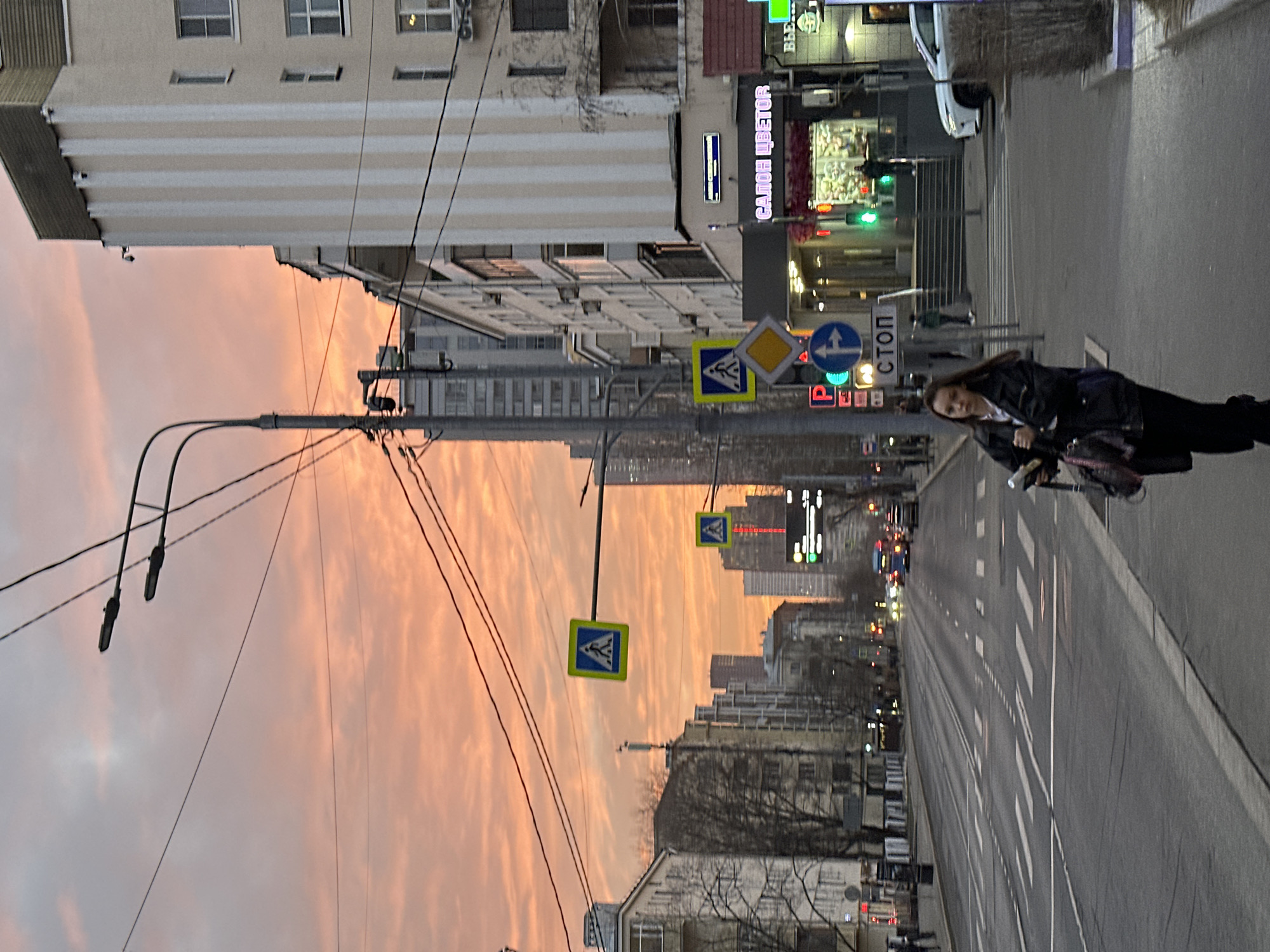 The image displays an urban street scene at dusk or dawn in Moscow, Russia, specifically on Bolshaya Yakimanka Street, looking northeast. The sky exhibits prominent orange and pink hues with scattered clouds.
In the foreground, slightly right of center, a young female pedestrian with dark, shoulder-length hair is walking on the sidewalk. She wears a dark jacket, a light-colored shirt, and dark pants, carrying a dark bag in her left hand and possibly papers in her right.
To the immediate right, a multi-story building (at least seven visible floors) with a cream-colored facade and vertical white stripes is present. Its ground floor features an illuminated sign reading "САЛОН ЦВЕТОВ" (Flower Salon) above a recessed entrance with stairs leading down. A white SUV is parked on the sidewalk near this entrance. The building is identifiable as 27/29 Bolshaya Yakimanka Street.
The wide asphalt road in the midground has multiple lanes marked with white dashed and solid lines, including a pedestrian crossing. Overhead, numerous power lines crisscross the sky, supported by a tall, dark street light pole equipped with multiple luminaires. Various traffic signs are visible: two yellow square signs depicting a walking person (pedestrian crossing), a yellow diamond-shaped sign, a blue circular sign with a white arrow pointing left (indicating a mandatory turn or restriction), and a white rectangular sign with "СТОП" (STOP) in blue Russian letters below the blue arrow sign. A traffic light shows a green signal for vehicle traffic.
In the background, multi-story buildings line both sides of the street, with bare trees obscuring some structures on the left. Distant modern skyscrapers are visible against the horizon. Several vehicles, including a blue bus and cars with illuminated headlights, are present on the road. Indistinct pedestrian figures are also visible on the sidewalks further down the street.