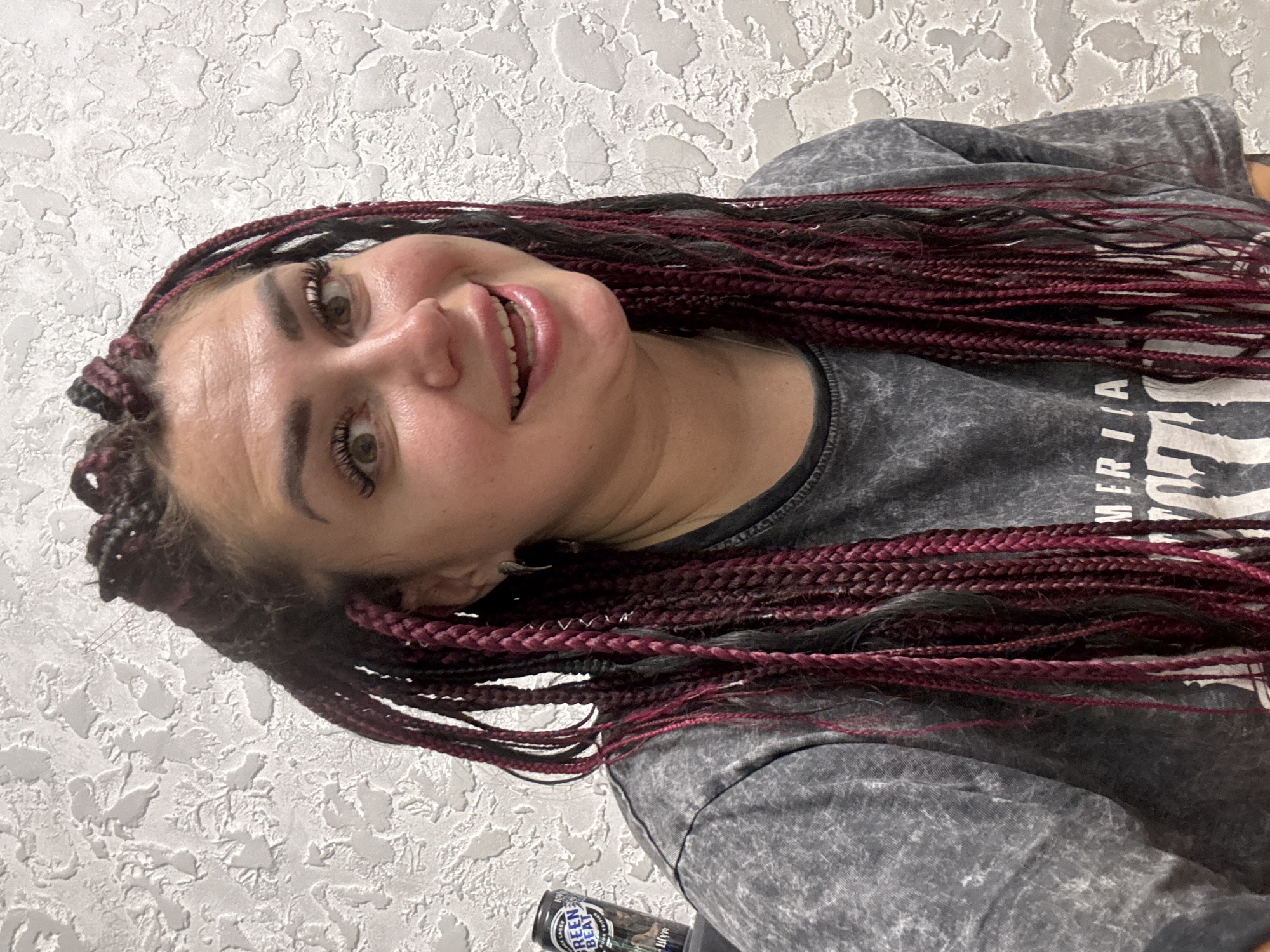 A close-up, medium shot shows a woman with dark hair styled in braids with magenta highlights. She is looking at the camera with a wide smile, her eyes are open and her eyebrows are raised. She is wearing a grey t-shirt. To her left, a can of "GREEN BEAT" black lager is visible. The background is a textured, light-colored wall. The image was taken indoors, and no specific location in Tolyatti, Russia can be identified.