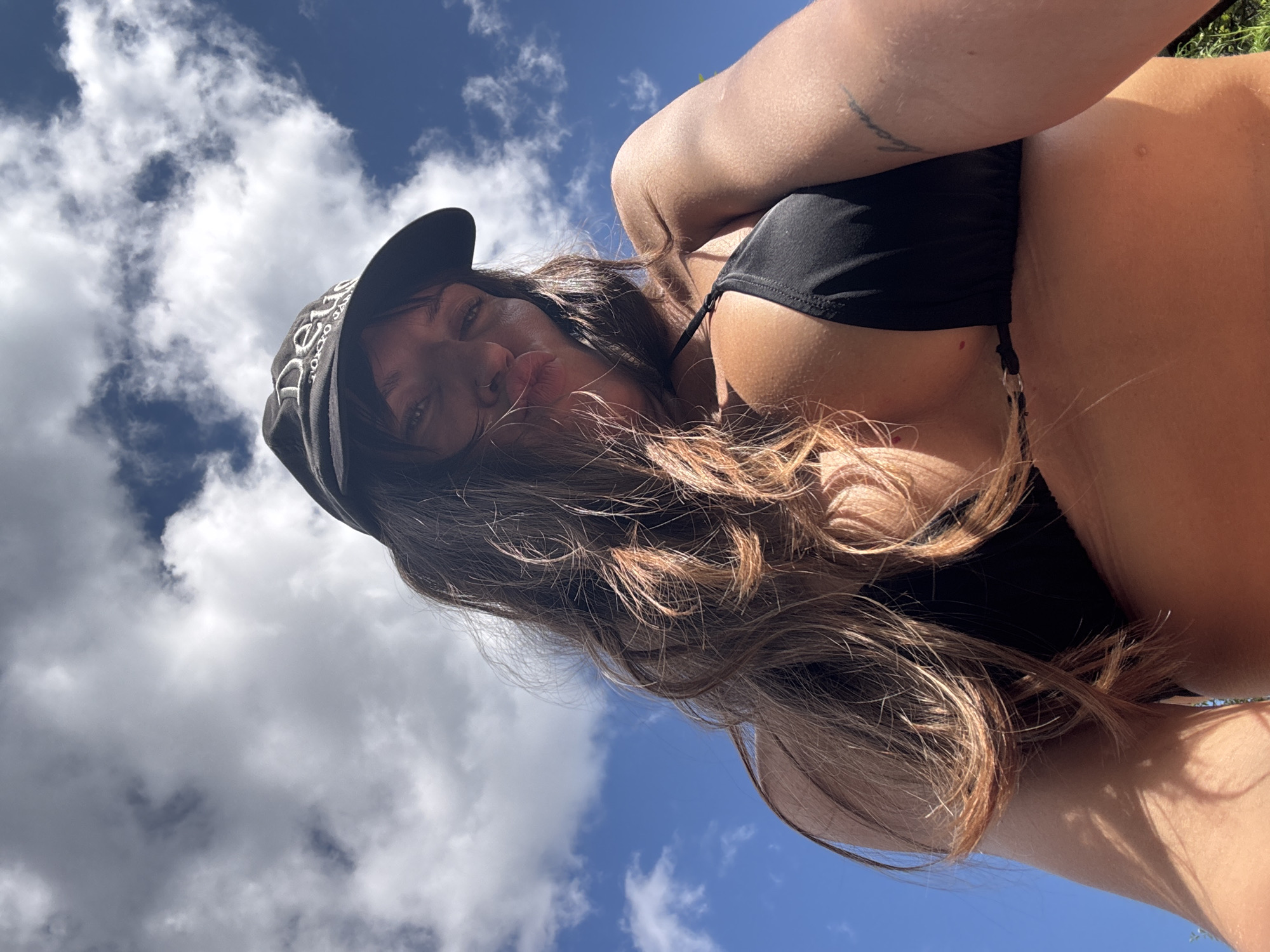 A low-angle, close-up, point-of-view shot shows a woman wearing a black bikini top and a black baseball cap with "Deus" and "Tokyo Japan" embroidered on it. She is looking up at the camera with her lips pursed. Her long, brown hair is blowing in the wind, and a tattoo is visible on her upper right arm. The background is a bright blue sky with scattered white clouds. The image appears to have been taken outdoors on a sunny day. There is no discernible information in the image to suggest a specific location in Krasnaya Presnya SNT, Russia.