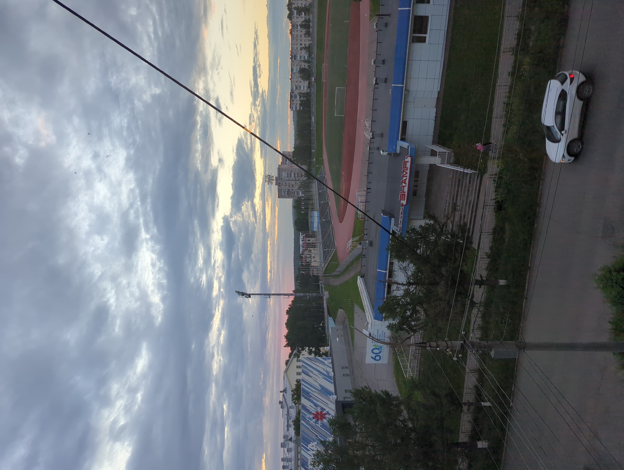 The image captures a twilight scene in Votkinsk, Russia, featuring a view of the "Zenit" stadium complex and surrounding urban environment. In the foreground, a white SUV is driving on a paved road, with a prominent utility pole and overhead wires. Behind the road, a paved walkway is bordered by a grassy area and trees. The "Zenit" stadium is visible, characterized by a red running track, a green field, and spectator seating. Adjacent to the stadium is a building with blue and white siding, bearing the partially obscured name "SHAMA". Further in the background, multi-story residential buildings and other urban structures can be seen, set against a cloudy sky. The sky displays subtle hues of pink and orange, indicative of sunset or sunrise.