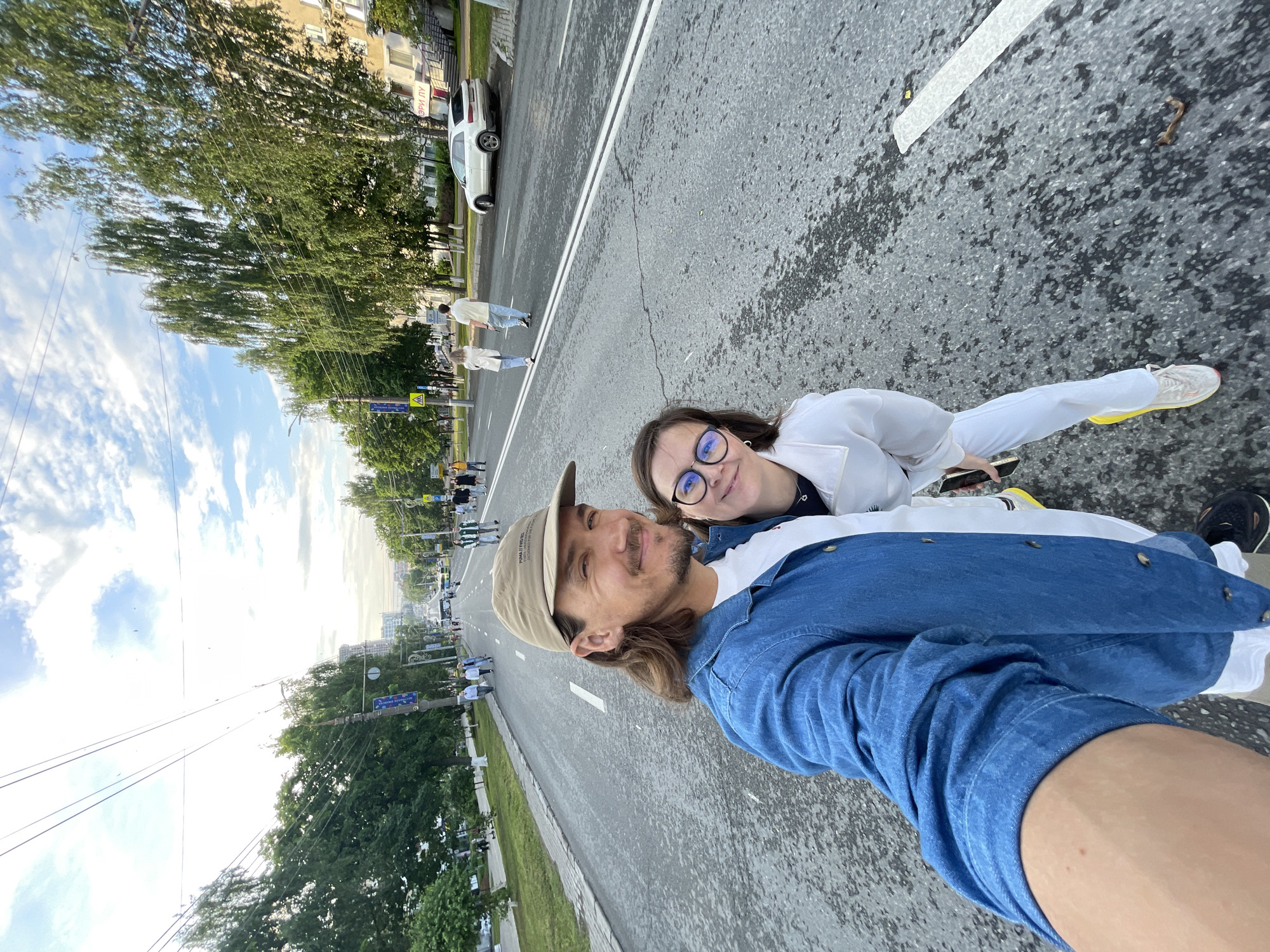 In a street scene in Unknown, Unknown, a man and a woman are taking a selfie. The man in the foreground wears a denim jacket and a beige baseball cap. He has long, brown hair and a mustache. The woman next to him has short brown hair, glasses, and wears a white jacket. They are positioned on a paved road with visible lane markings. In the background, there are trees lining the street, buildings, and several people walking. A white car is parked on the side of the road. The sky above is partly cloudy.