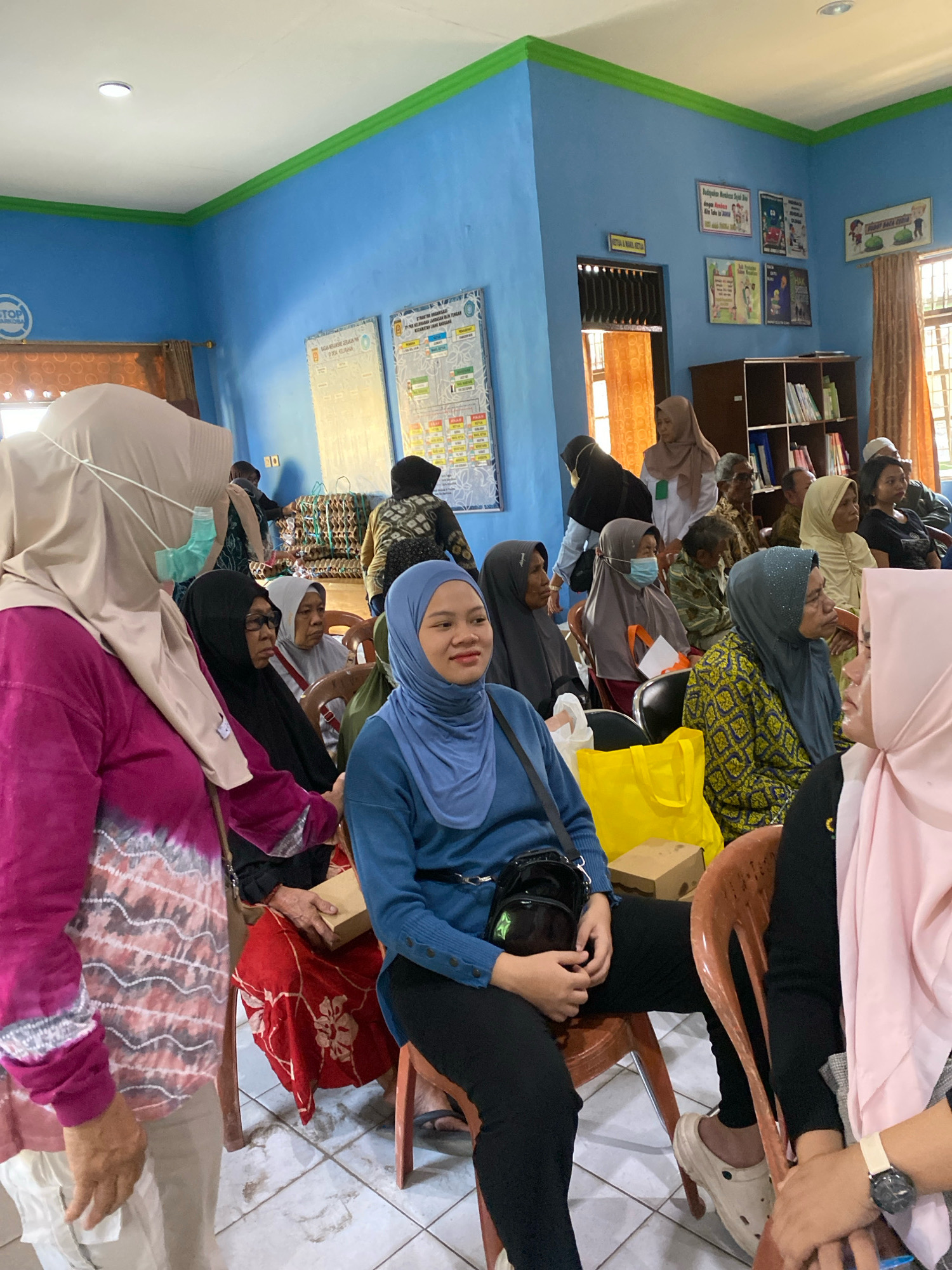 The image captures an indoor gathering, likely a community event or meeting, within a room painted in vibrant blue and green. Several individuals, predominantly women wearing headscarves, are seated on chairs, facing a central point, suggesting an ongoing presentation or discussion. The room's environment is characterized by posters and charts displayed on the walls, a bookshelf filled with books, and large windows providing natural light. The presence of a "STOP NARKOBA" sign on the wall indicates a possible anti-drug campaign or awareness program related to the event. A few individuals are wearing masks, indicating public health precautions. The overall atmosphere appears calm and attentive, with the participants engaged in the activity. The location is identified as Banjarbaru, Indonesia, further contextualizing the cultural and social setting of the scene.