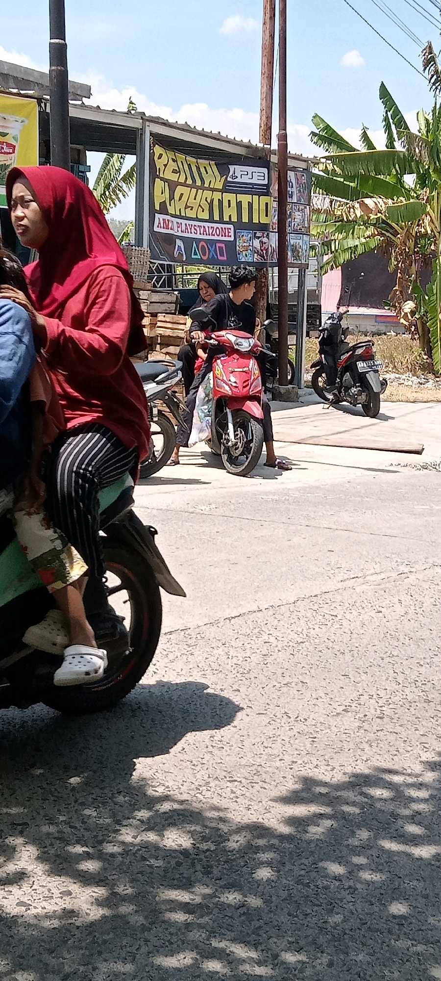In Desa Karangmojo, Indonesia, a group of people are on motorcycles. In the foreground, a woman wearing a red hijab and striped pants sits on the back of a green and black motorcycle. She is wearing white clogs. To her right, a red motorcycle is being ridden by a young man in a black shirt, with a woman in a black hijab behind him. A second black motorcycle is parked behind them, and another motorcycle is visible further in the background. Behind the people and motorcycles, a banner advertises "RENTAL PLAYSTATION" and "PLAYSTAIZONE" with PlayStation logos. The scene appears to be outdoors on a paved surface under a bright, sunny sky.