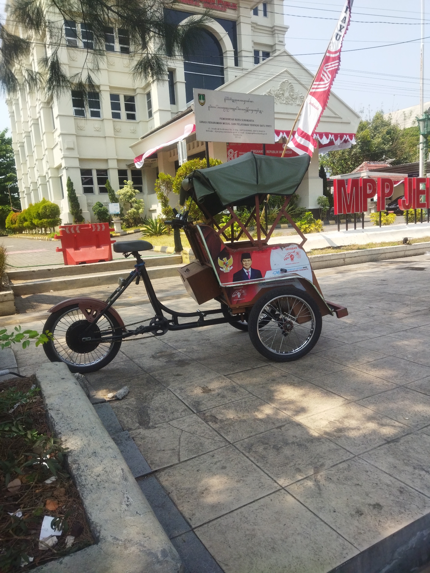 In the city of Surakarta, Indonesia, a three-wheeled vehicle, possibly a becak or pedicab, is parked on a tiled pedestrian area. The vehicle is dark-colored with a green canopy and has a picture of a man in a suit on its side. Behind the vehicle, a light-colored, multi-story building stands with several windows and an arched entryway. Red and white Indonesian flags are visible, one of which is partially obscuring the building's facade. In front of the building, large red letters spell out "MPP JE". Several trees and bushes are present in the scene, and the ground is paved with square tiles. Red traffic barriers are also visible in the background. The scene appears to be a public area in front of a government building.
