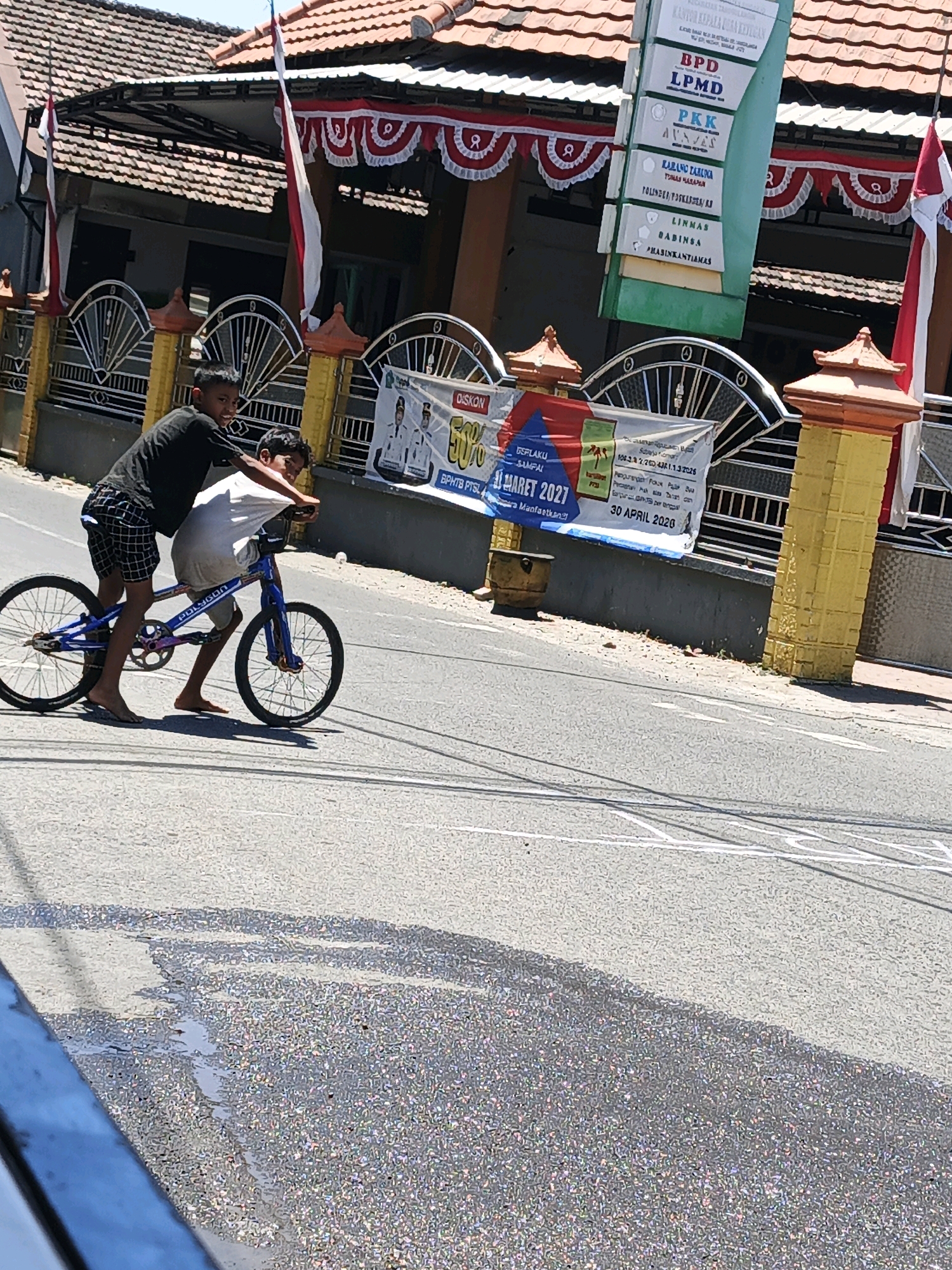 In the city of Ketegan, Indonesia, two young boys are captured in a moment of playful interaction on a street under a bright, sunny sky. The foreground is dominated by an asphalt road, showing signs of recent moisture with scattered puddles reflecting the sunlight. The midground features a blue bicycle with two young males aboard. One boy, wearing a black t-shirt and checkered shorts, is positioned at the front, appearing to steer or balance the bike. The second boy, clad in a white t-shirt, is seated behind the first, his arms around him, suggesting a shared ride. Both boys are barefoot. The bicycle itself is a BMX style with visible chain and pedals.
Behind the cyclists, a paved sidewalk or pathway is visible, leading to a building with a red-tiled roof. The building features an awning supported by dark pillars and decorated with festive red and white bunting. A yellow pillar with decorative detailing stands prominently on the right side of the frame. A tall, green signpost with various labels, including "BPD," "LPMD," and "PKK," is tilted at an angle in front of the building. The background consists of the building's facade and the clear blue sky. A banner with Indonesian text and graphics, possibly advertising an event or service, is partially visible, hanging between a gate and the yellow pillar. The overall scene depicts a casual, outdoor activity in a residential or public area.