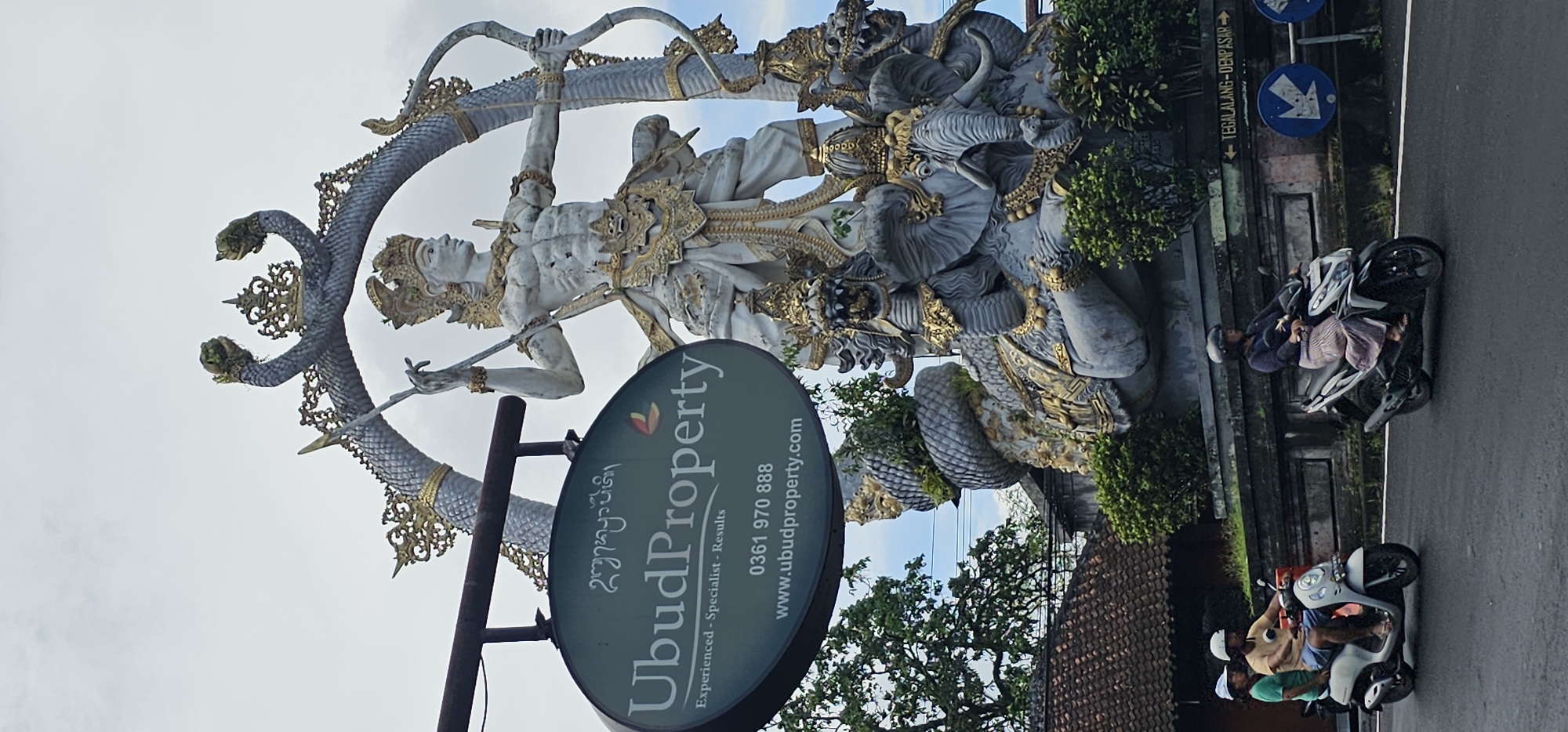 The image shows a street scene in the Ubud District of Indonesia. A large statue of a male figure is prominently displayed, surrounded by a seven-headed snake Naga and other decorative elements. A sign for "Ubud Property," a real estate company, stands in the foreground. Two motorbikes are parked or driving along the road, one with two riders and another with one. A street sign indicates the direction to "TEGALALANG-DENPASAR." The environment includes buildings, trees, and a partly cloudy sky.