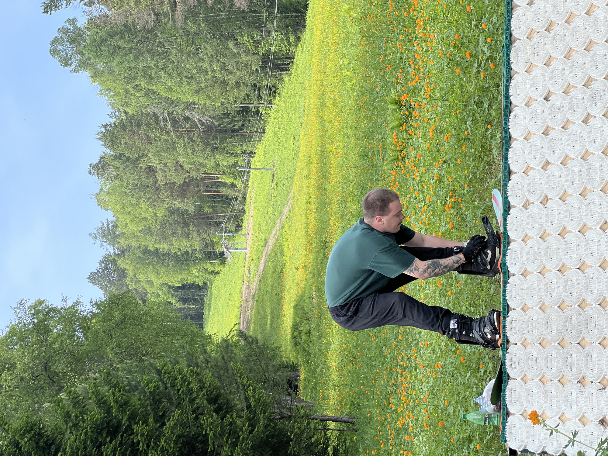 In an outdoor setting, a person with tattoos on their arm is seen bending over to adjust their snowboard bindings. They are wearing a green t-shirt and black pants. The snowboard is positioned on a mat with a white, circular, textured surface. In the background, there is a lush green slope covered in small orange flowers, with a line of trees bordering the top. A ski lift structure is visible on the slope. A plastic water bottle is also present near the snowboard. The sky is clear and blue.