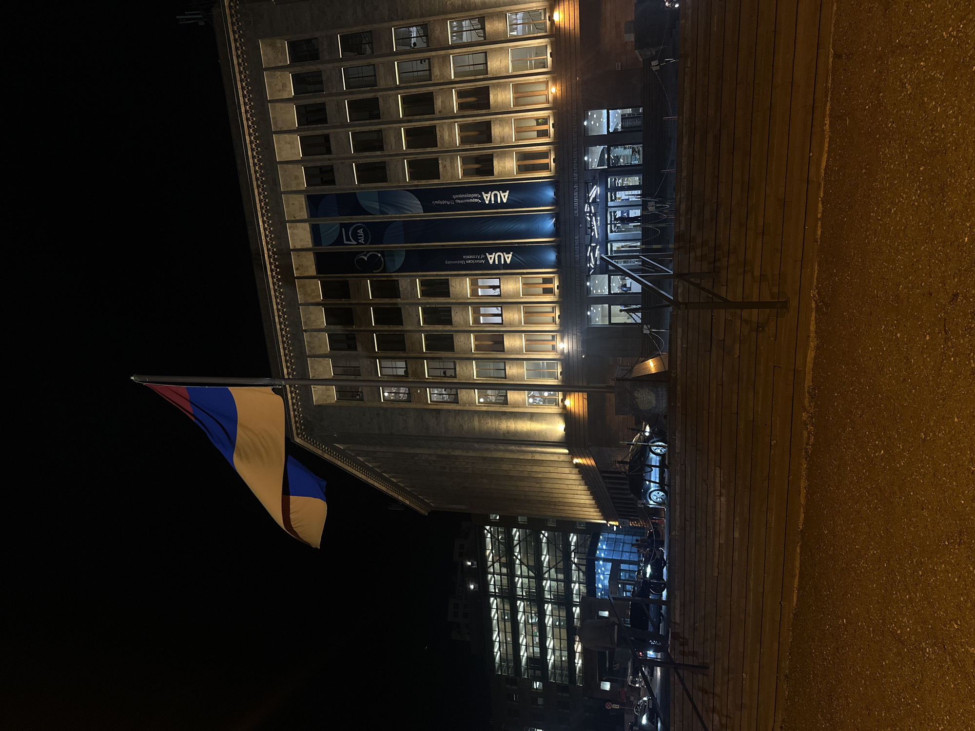 At night in Yerevan, Armenia, a flag with red, blue, and orange stripes hangs from a pole near the American University of Armenia (AUA). The building is multi-storied and features rows of windows. Blue banners bearing the "AUA" logo are visible on the building's facade. The foreground shows a paved area. Cars are parked on the street nearby. The building has many lit windows.