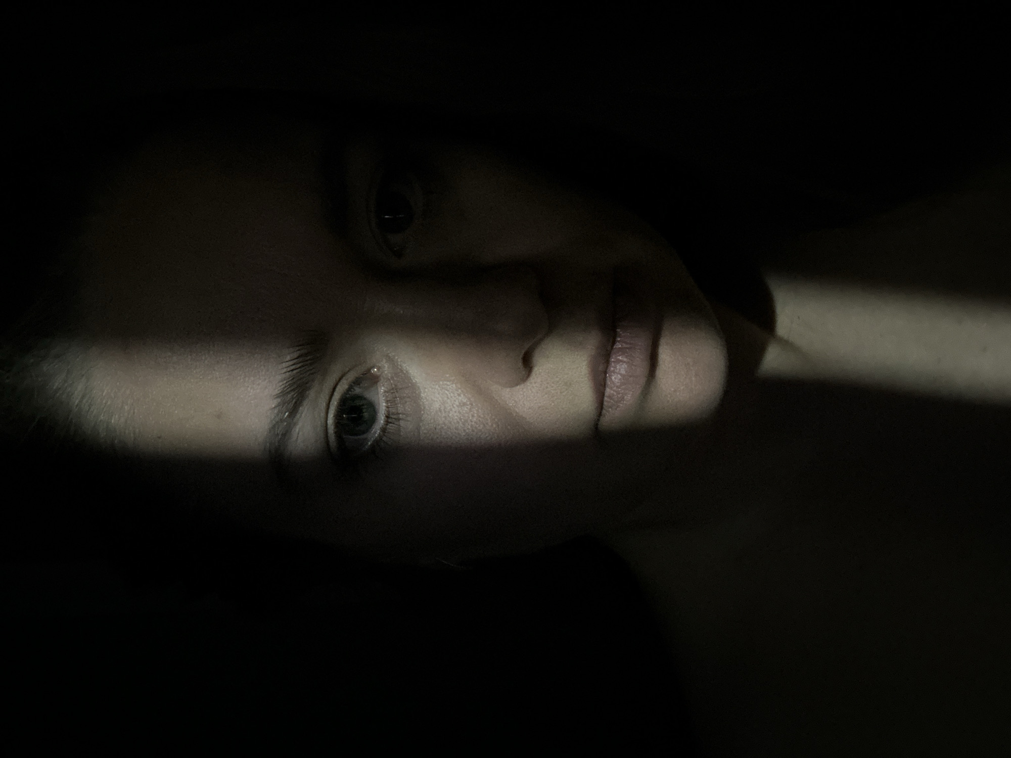 The image is a close-up of a person's face, dramatically lit with a stark line of light and shadow. The person is lying down, with their face angled towards the viewer. One eye is fully visible, while the other is partially obscured by shadow. The line of light crosses the face diagonally, highlighting part of the nose and lips. The background is dark and indistinct. The image provides no information about a location in the city of Chaikovsky, Russia.