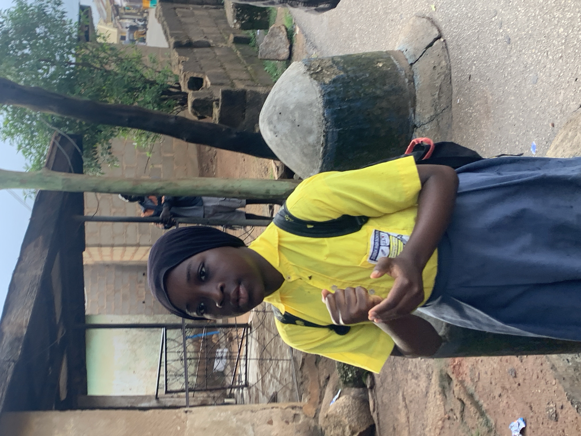 The image shows a young person wearing a bright yellow school uniform and a black headscarf, with a dark backpack on. The person is looking towards the camera with a neutral expression. They are standing in what appears to be an outdoor, possibly urban, environment. In the background, there are brick buildings, some overgrown with vegetation, and a large, dark, somewhat conical object on the ground. A tree branch is prominently visible in the foreground, partially obscuring the scene. The ground is paved with what looks like asphalt or concrete. There are some rocks and debris scattered around. The overall lighting suggests it is daytime, possibly overcast or late afternoon given the soft light. The attire of the person and the visible buildings hint at a setting in a place like Jos, Nigeria, likely on school grounds or a street near a school. A small patch of blue and white material, possibly litter, is visible on the ground. There is text visible on a yellow patch on the person's shirt, but it is too blurry to read.