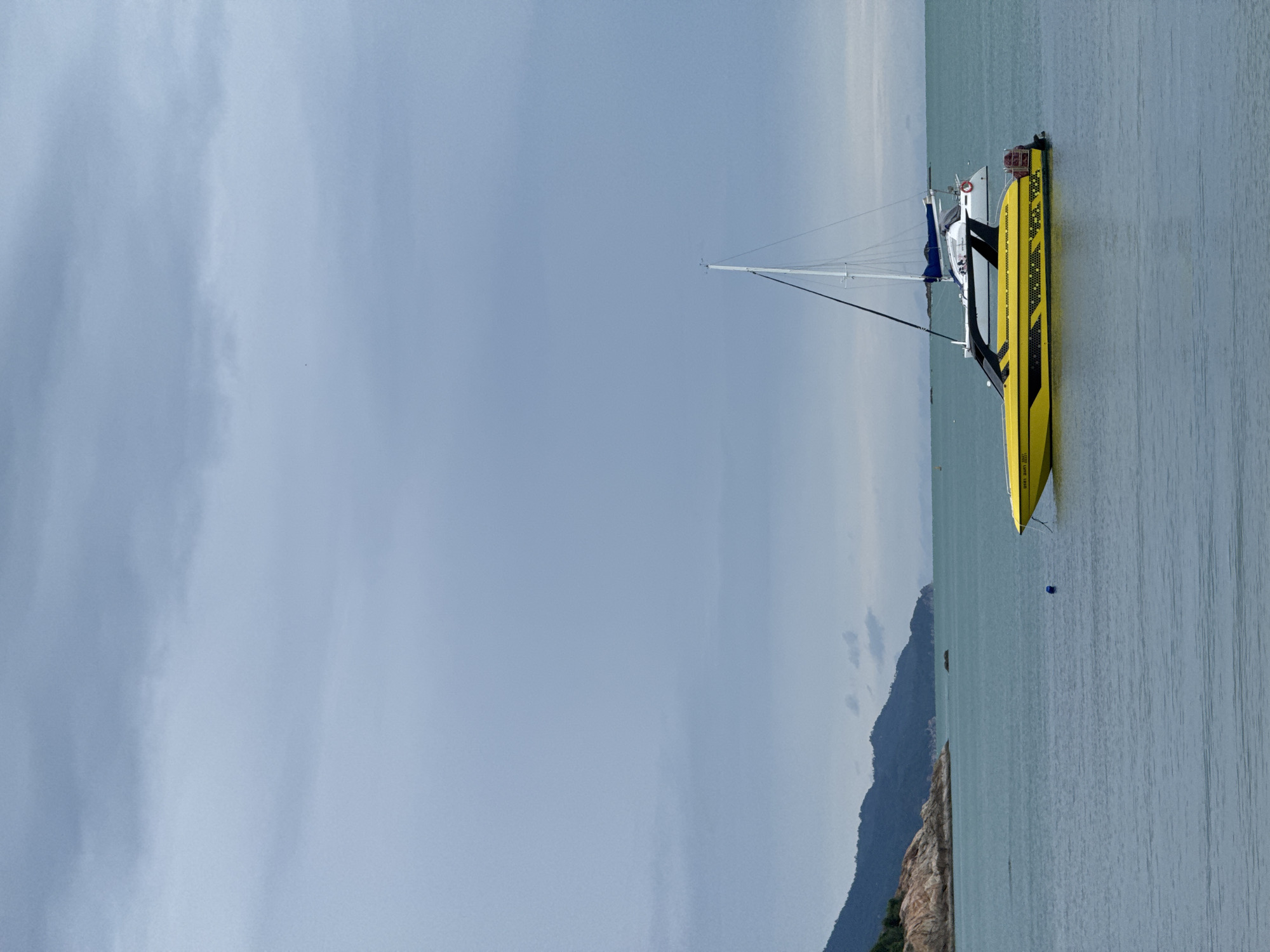 A yellow speedboat is anchored in the ocean, with a white sailboat in the background. The water is calm and a light blue, and the sky is overcast with gray clouds. In the distance, there is a mountainous island with green trees and rocky shores. The scene appears to be in a tropical or subtropical location.