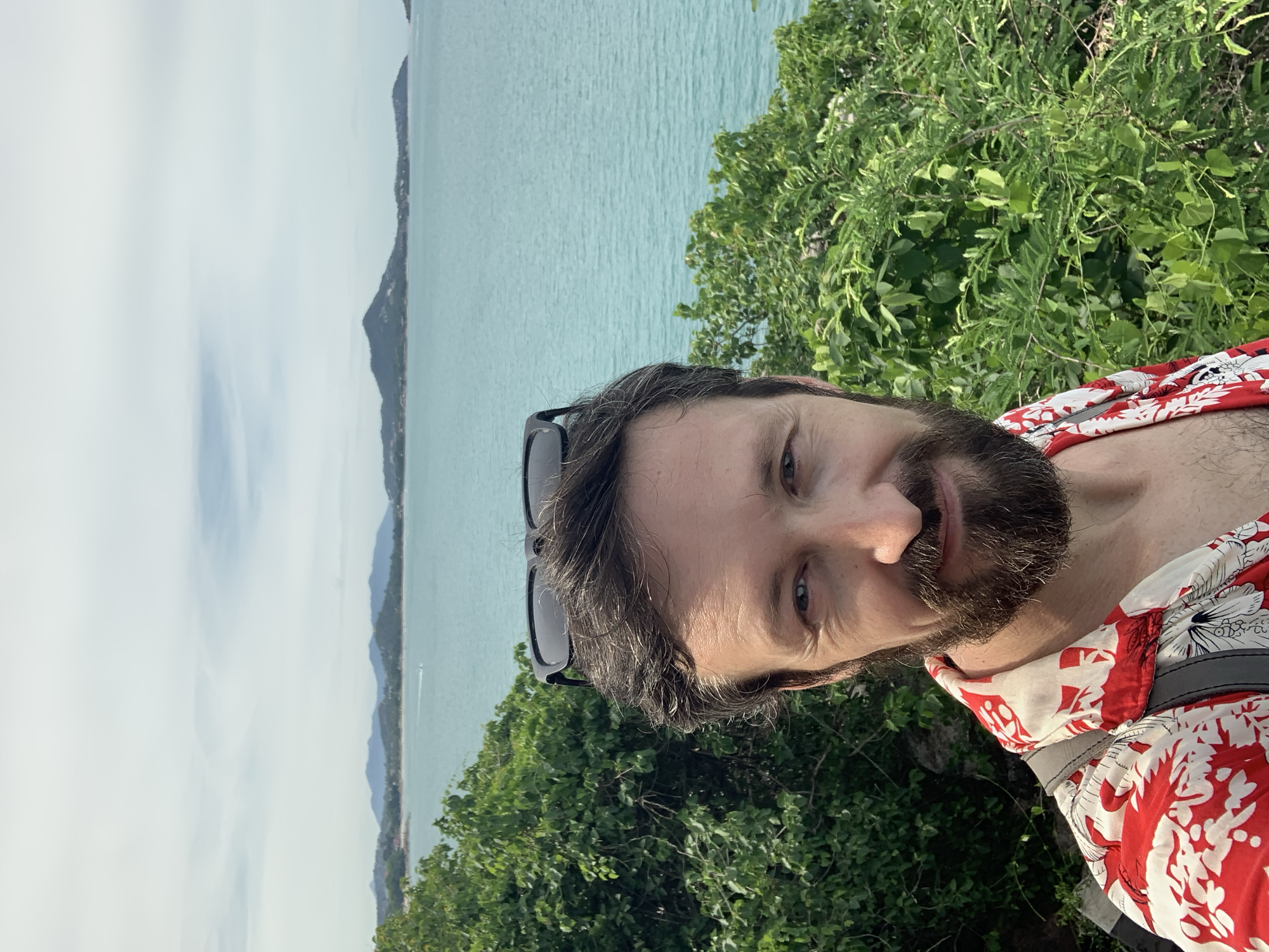 A medium shot selfie shows a Caucasian man with dark hair and beard against a backdrop of lush green foliage and the turquoise sea. He is wearing a red and white patterned short-sleeved shirt and has sunglasses perched on top of his head. The man's face is turned towards the camera, and he has a slight smile. In the background, islands and mountains are visible across the calm ocean under a partly cloudy sky. The scene suggests a tropical location, possibly Baan Chaweng Noi, Thailand, known for its scenic beauty and coastal landscapes.