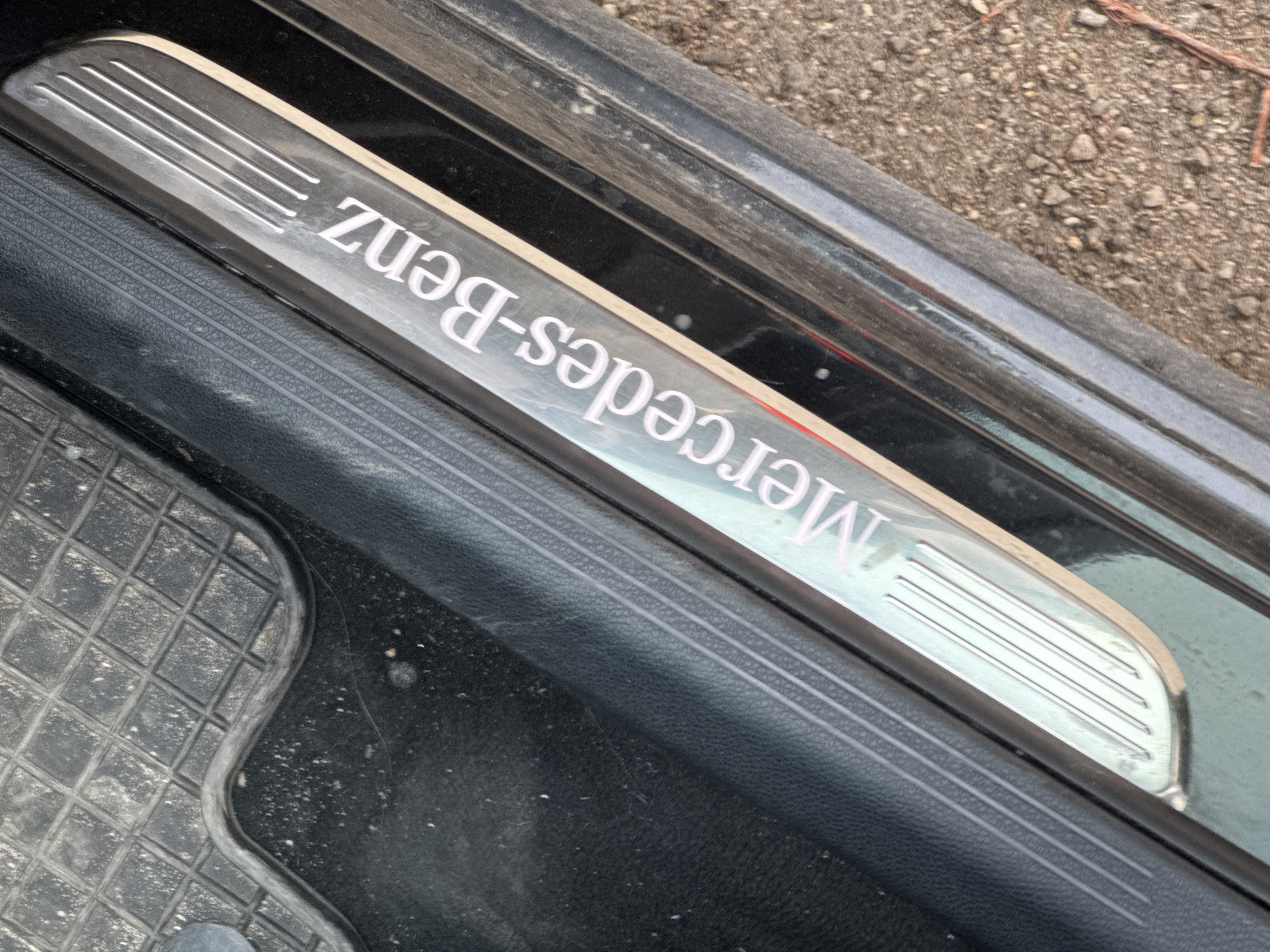 The image shows a close-up of a Mercedes-Benz door sill, likely inside a car. The sill is metal with the "Mercedes-Benz" logo inscribed. The door frame is black, with a rubberized seal. A part of a black, patterned floor mat is visible in the lower left corner. The environment outside the car is blurred and appears to be dirt or gravel. The city and country are not determinable from the image.