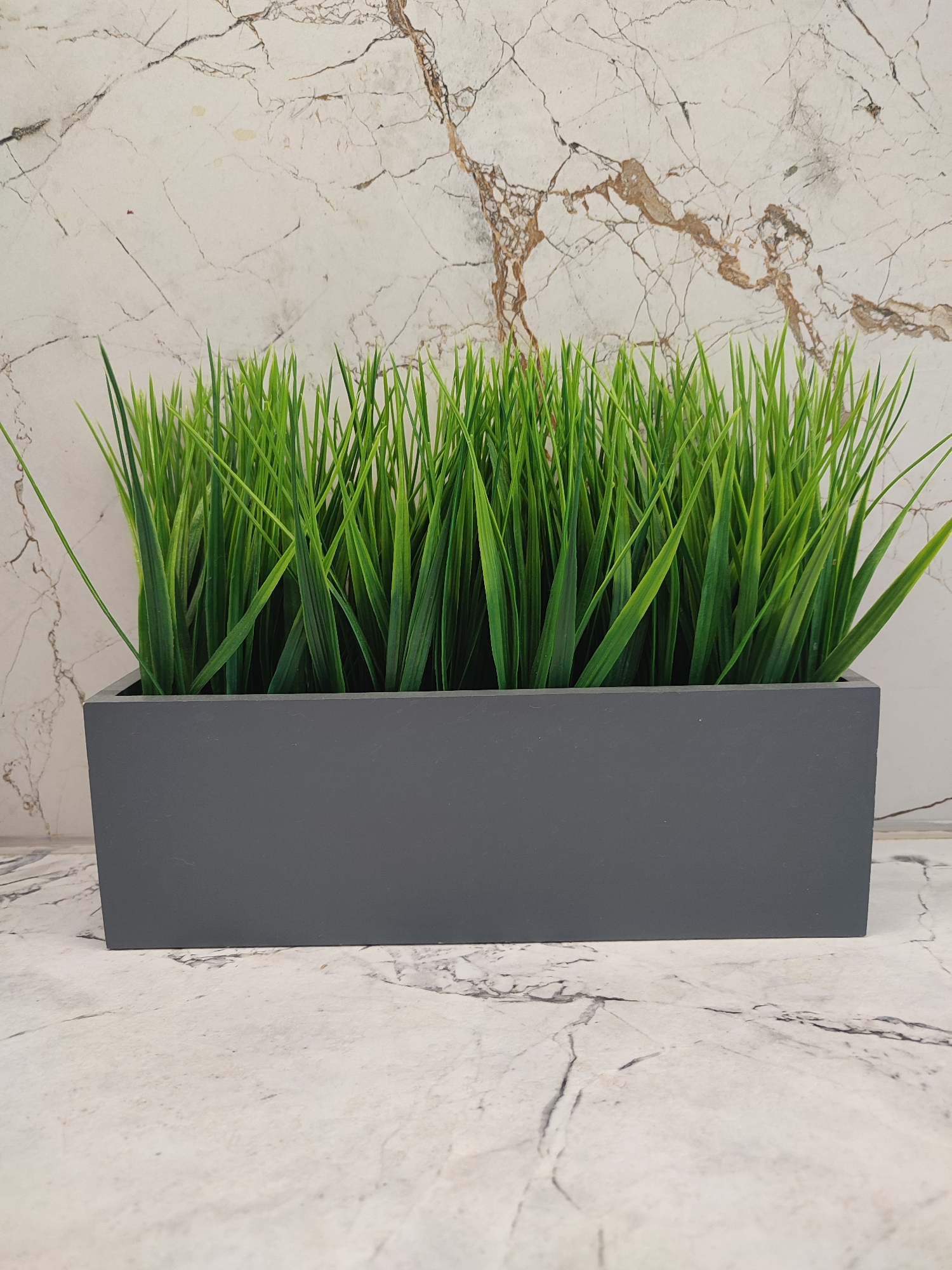 A rectangular, dark gray planter filled with artificial green grass sits on a white marbled surface. The planter is positioned in the foreground, centered in the frame. Behind the planter, the background is a wall with a similar white marble pattern, featuring brown veining. The lighting is even, and the image is taken from a straight-on, eye-level perspective. There are no people or animals present in the scene.