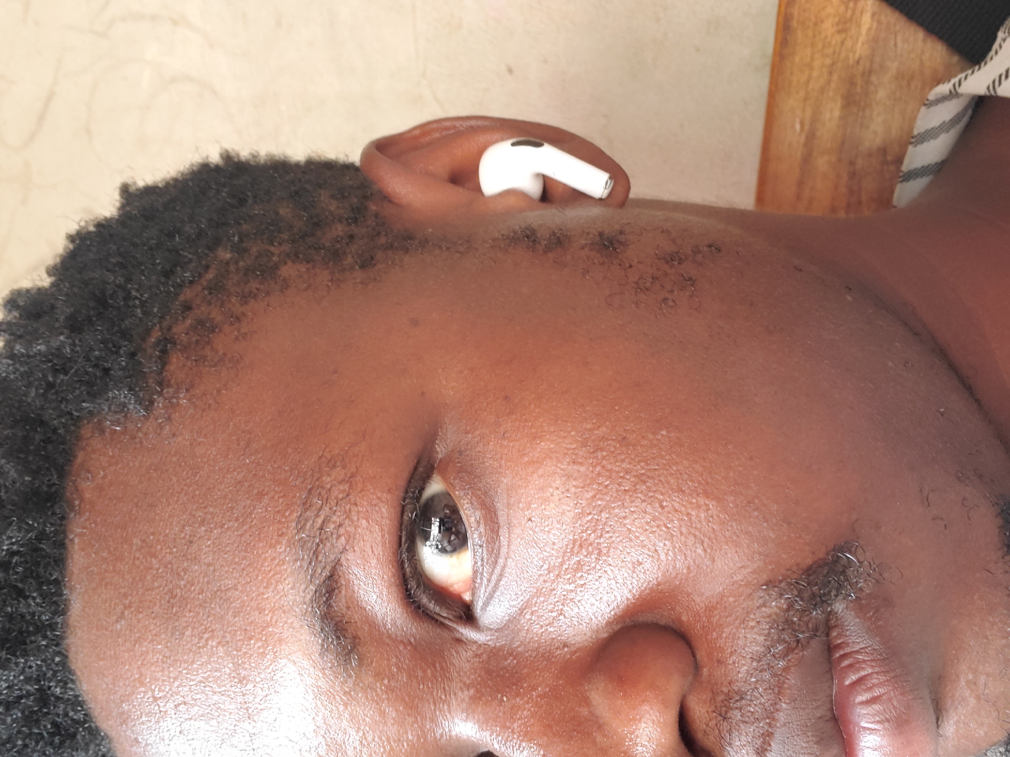 This is a close-up, portrait-style image of a person, likely male, with dark skin and short, dark, curly hair. The image is rotated 90 degrees clockwise, orienting the person's head sideways. A white earbud, specifically an Apple AirPods Pro, is visible in the person's left ear (viewer's right). The person's right eye is open and appears to be looking upwards relative to their head, with a neutral expression. Short, dark facial hair, including an eyebrow, a mustache, and stubble on the chin/jawline, is present.
The skin texture is clearly visible, showing pores and fine hair. In the background, to the upper right, a wooden surface can be seen, partially obscured by a striped fabric (black and white), which might be part of clothing, a pillow, or upholstery. The lighting is soft and even, suggesting an indoor setting without harsh direct light. There is no discernible specific activity beyond the individual being present, possibly resting or listening to audio. No text is visible in the image. The appearance of the individual is consistent with the provided location context of Mararaba, Nigeria.
