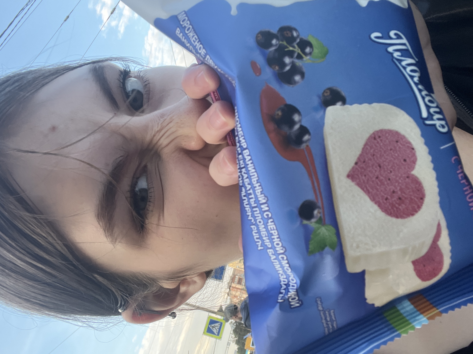 This image is a close-up, low-angle selfie taken outdoors in Irinovka, Russia, on a bright, sunny day. The primary subject is a person, largely depicted by their face—specifically, their left eye, nose, and forehead—and their right hand. The person's face is playfully scrunched up, and their eye looks directly into the camera. Dark hair frames their face, and an industrial piercing is visible on their left ear.
The person is holding a blue ice cream package, branded "Пломбир" (Plombir), close to their face. The packaging features illustrations of blackcurrants and a white ice cream bar with a prominent pink heart design. Visible text on the package includes "МОРОЖЕНОЕ ПЛ" (Ice Cream PL), "ПЛОМБИР ВАНИЛЬНЫЙ И С ЧЕРНОЙ СМОРОДИНОЙ" (Vanilla Plombir with Blackcurrant), "С ЧЕРНОЙ СМОРОДИНОЙ" (with Blackcurrant), and "ПЛОМБИР БАЛМҰЗДАҒЫ" (Plombir Ice Cream in Kazakh). Other partial words like "ВАНИЛЬ" (Vanilla) and "up" are also discernable.
The background shows a clear blue sky with some white clouds and overhead power lines. A yellow road sign, likely indicating a pedestrian crossing, is visible in the lower-left background, suggesting an urban or suburban setting. The scene captures a casual, lighthearted moment of the person posing with their ice cream.