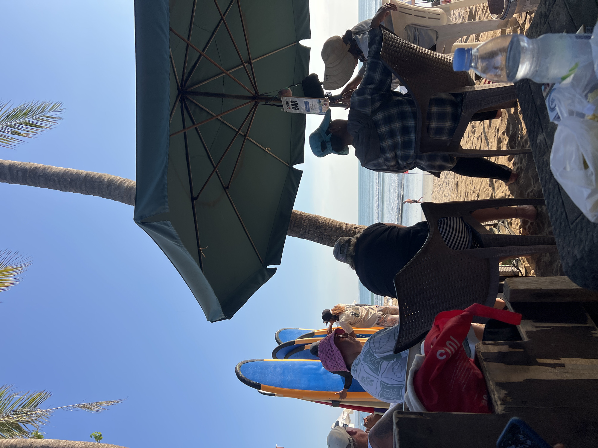 The image depicts a beach scene in Kuta, Indonesia, likely on the southwestern coast of Bali, known for its surf breaks. In the foreground, several people are seated at tables or chairs, with a large green umbrella providing shade. A plastic bottle of water and some bags are visible on a table. Behind them, a group of surfboards, primarily blue and yellow, are propped up. Further back, people are interacting near the surfboards, and the ocean with gentle waves is visible in the background. The sky is clear and blue, and a palm tree trunk is present on the right side of the frame. The lighting suggests it is daytime, possibly late afternoon.